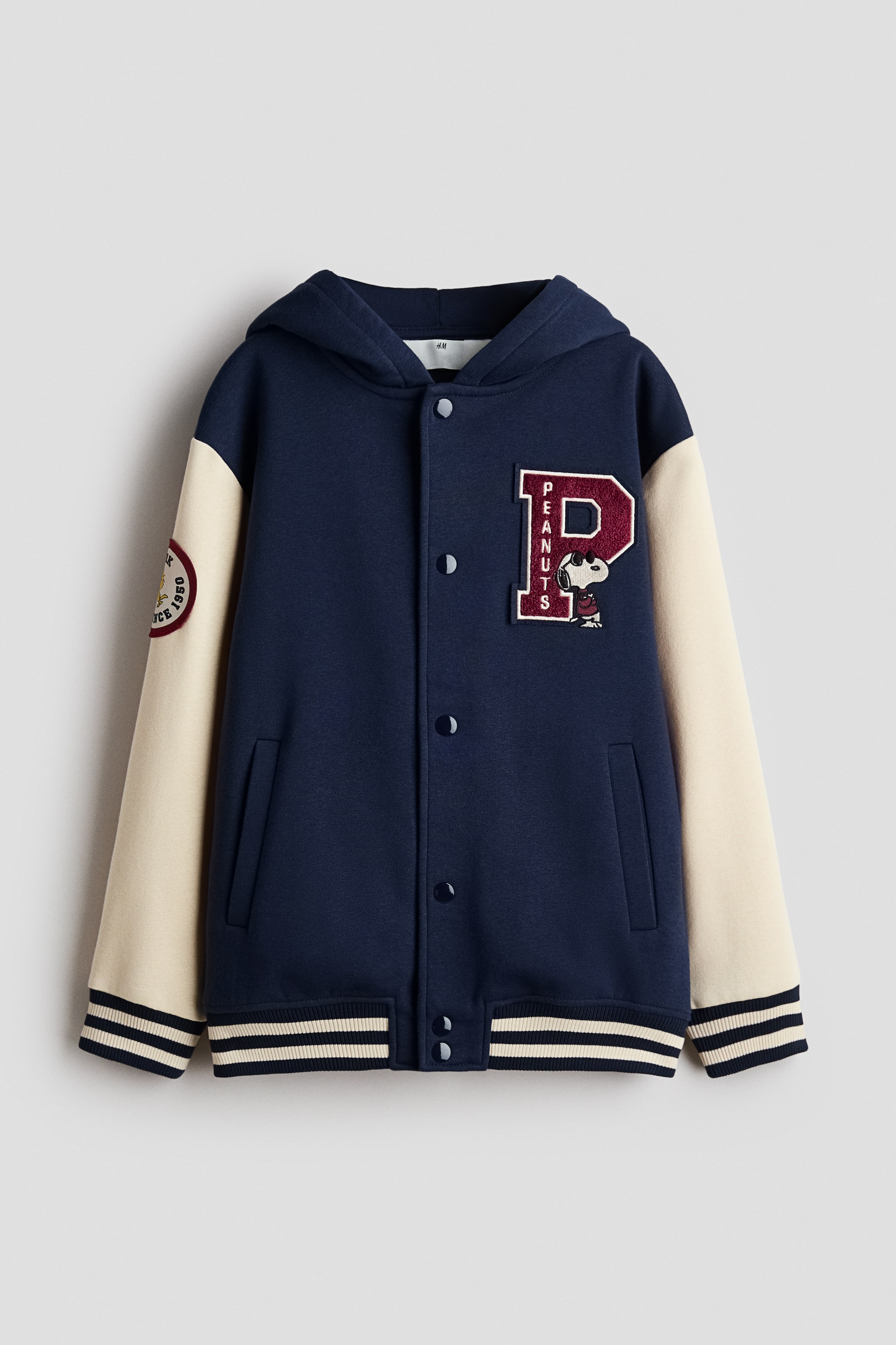 View larger image: A hooded varsity jacket features a navy blue body with cream-colored sleeves, facing forward. It showcases a dark red 'P' patch with 'PEANUTS' text and a Snoopy graphic on the left chest, a circular Snoopy patch on the left sleeve, a button-up front, angled welt pockets, and striped ribbed cuffs and hem.