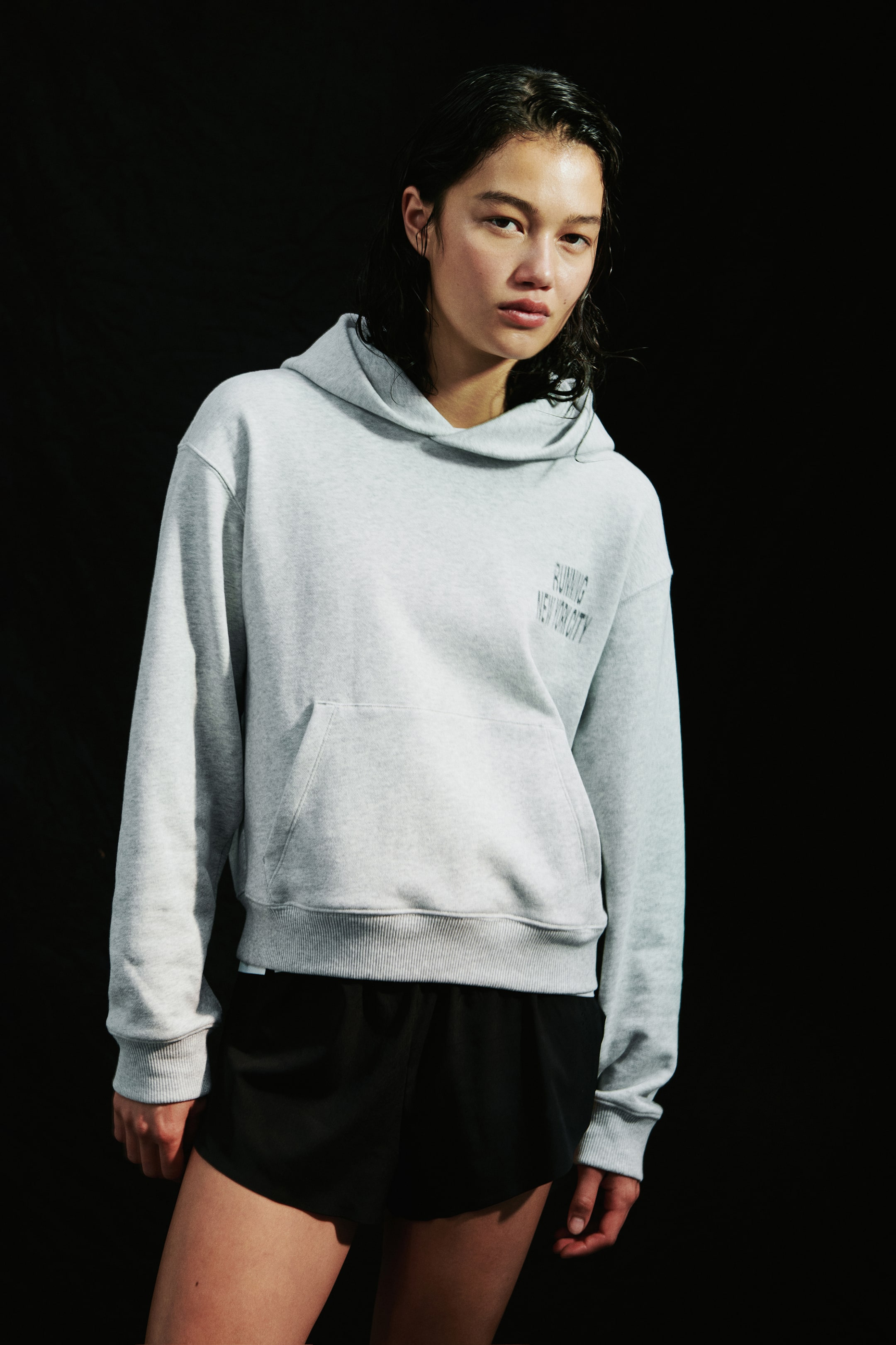 View larger image: A light gray hoodie featuring a small black text graphic on the left chest is being worn by the model. The hoodie includes a front kangaroo pocket, ribbed cuffs, and a ribbed hem, and is paired with black athletic shorts.