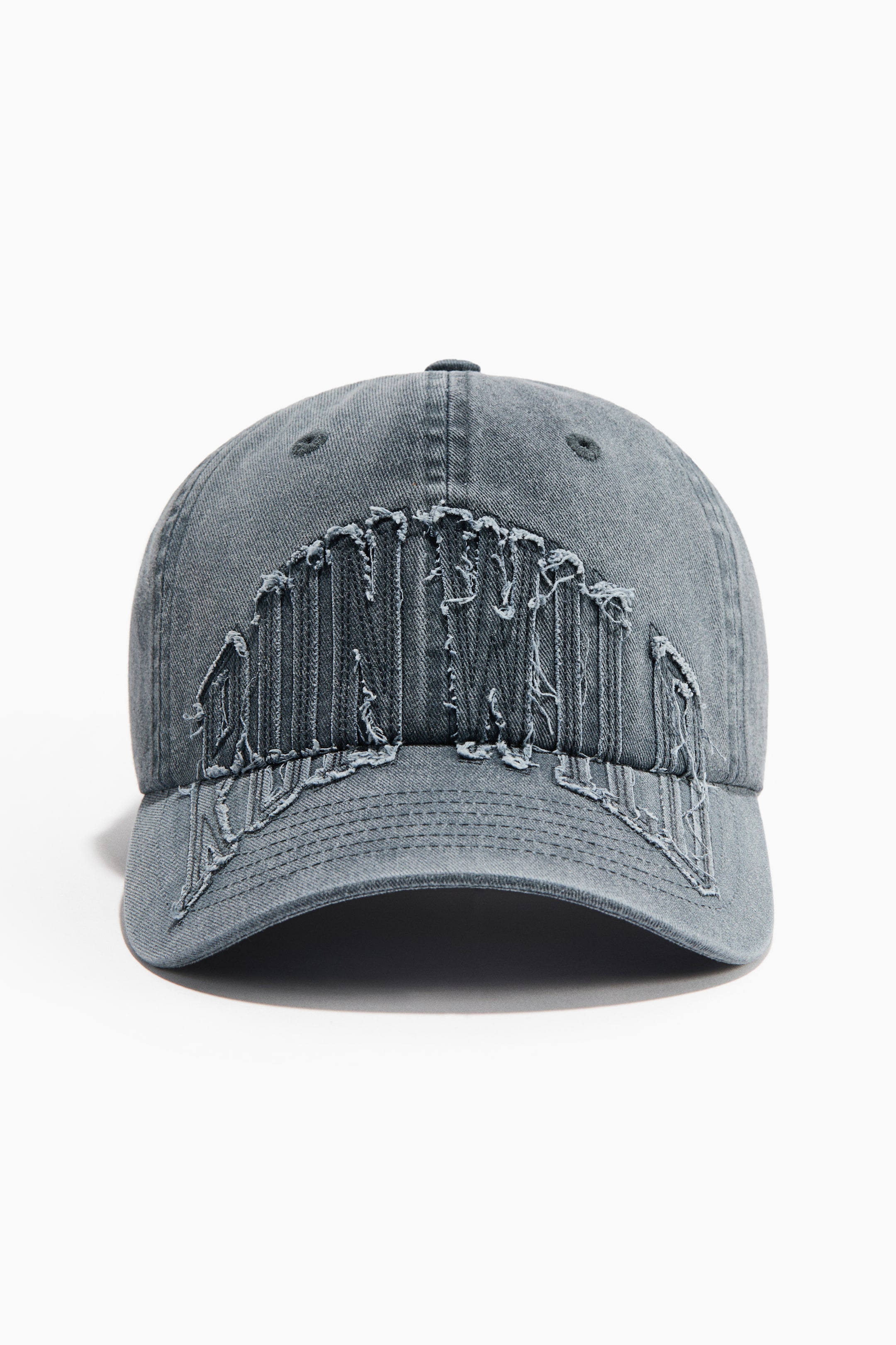 View larger image: A cotton twill cap is presenting its front view, featuring a large, distressed, frayed fabric design across the front crown and brim. The cap is a faded dark grey with a curved brim.