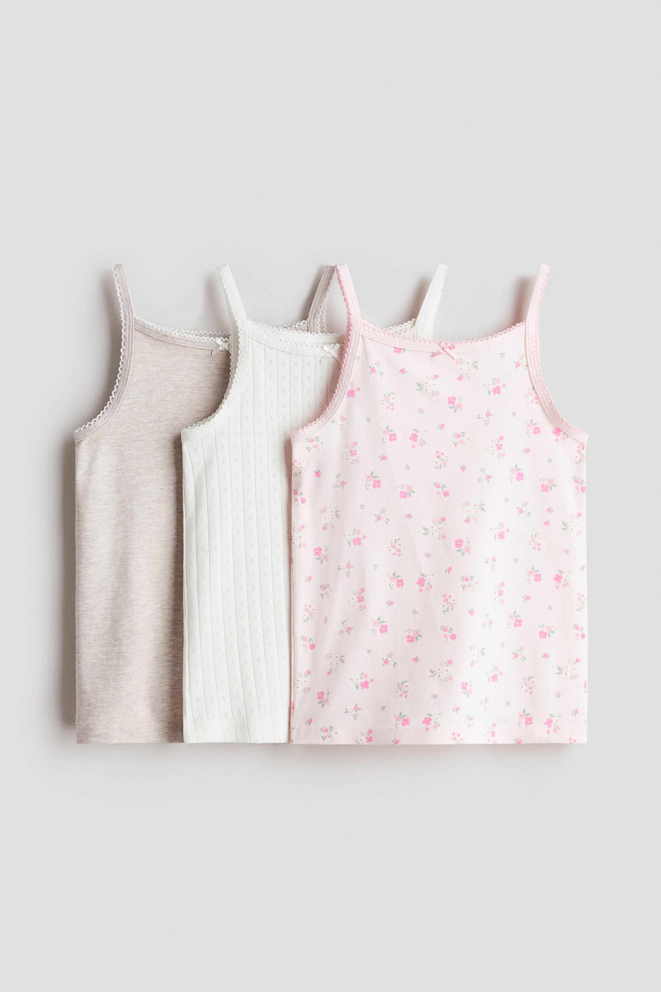 View larger image: "Three front-facing cotton jersey tops showcase picot trims along their square necklines and narrow shoulder straps, each adorned with a decorative satin bow. The set comprises a solid light beige melange top, a white top with a subtle ribbed texture, and a light pink top with a floral print."