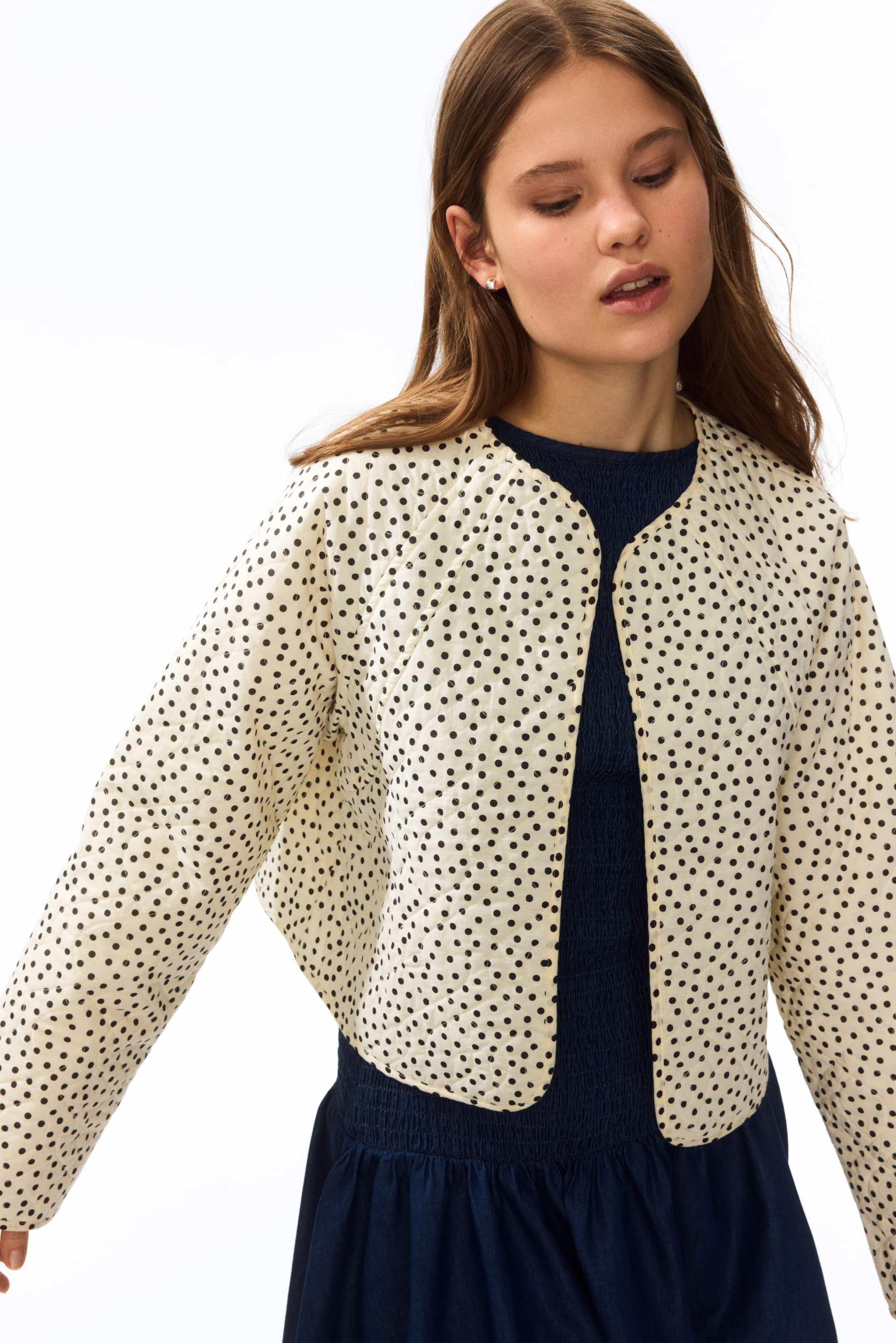 Agrandir l'image: La veste matelassée crème, ornée de pois noirs, présente une longueur raccourcie, des manches longues et une ouverture frontale avec un col rond. Elle est portée sur un vêtement bleu foncé et est légèrement orientée vers la droite du spectateur.