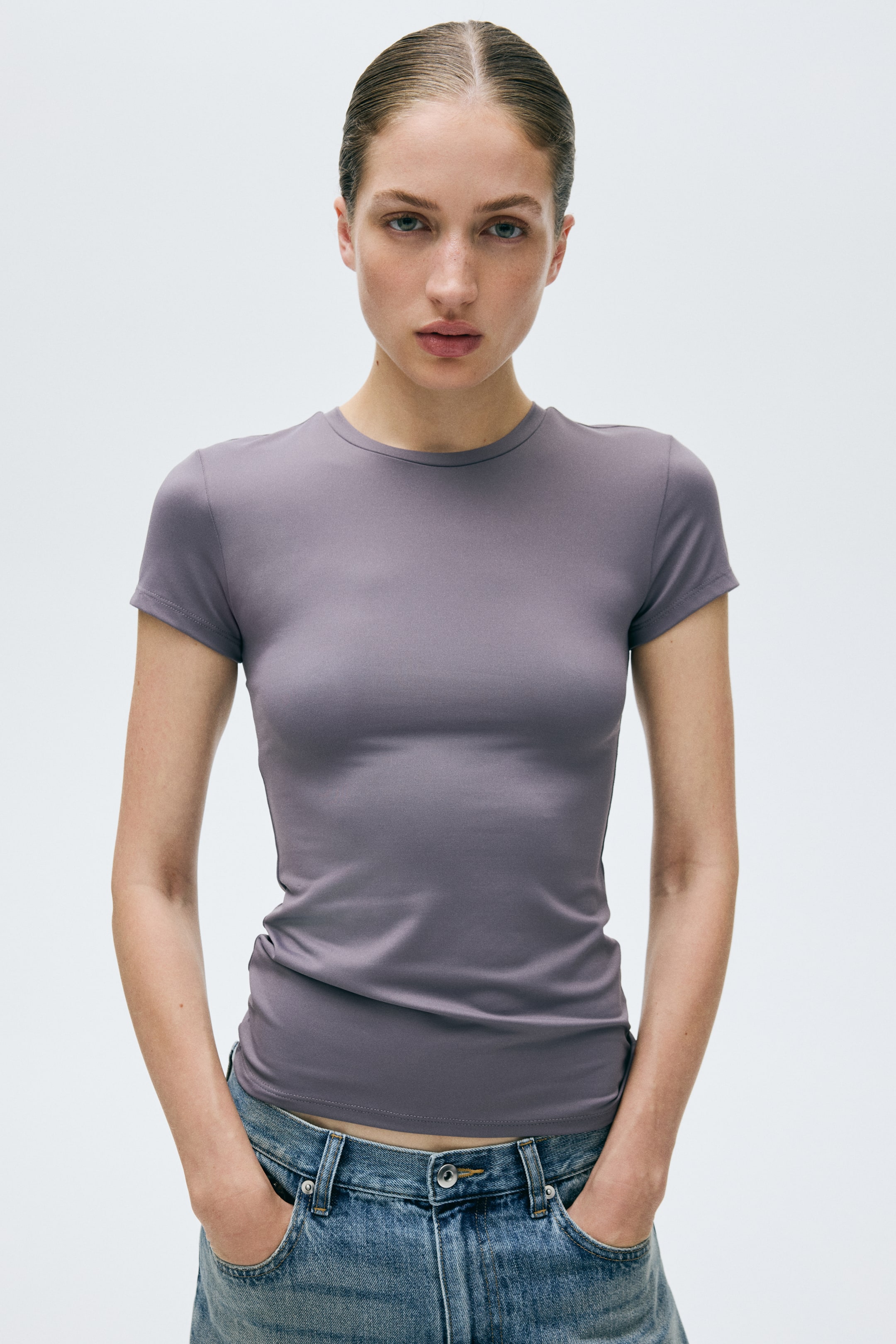 Visualizza immagine più grande: Una t-shirt slim-fit color viola tenue viene presentata frontalmente, con scollo a girocollo, maniche corte, orlo dritto che si ferma appena sopra la vita e una silhouette liscia e aderente.