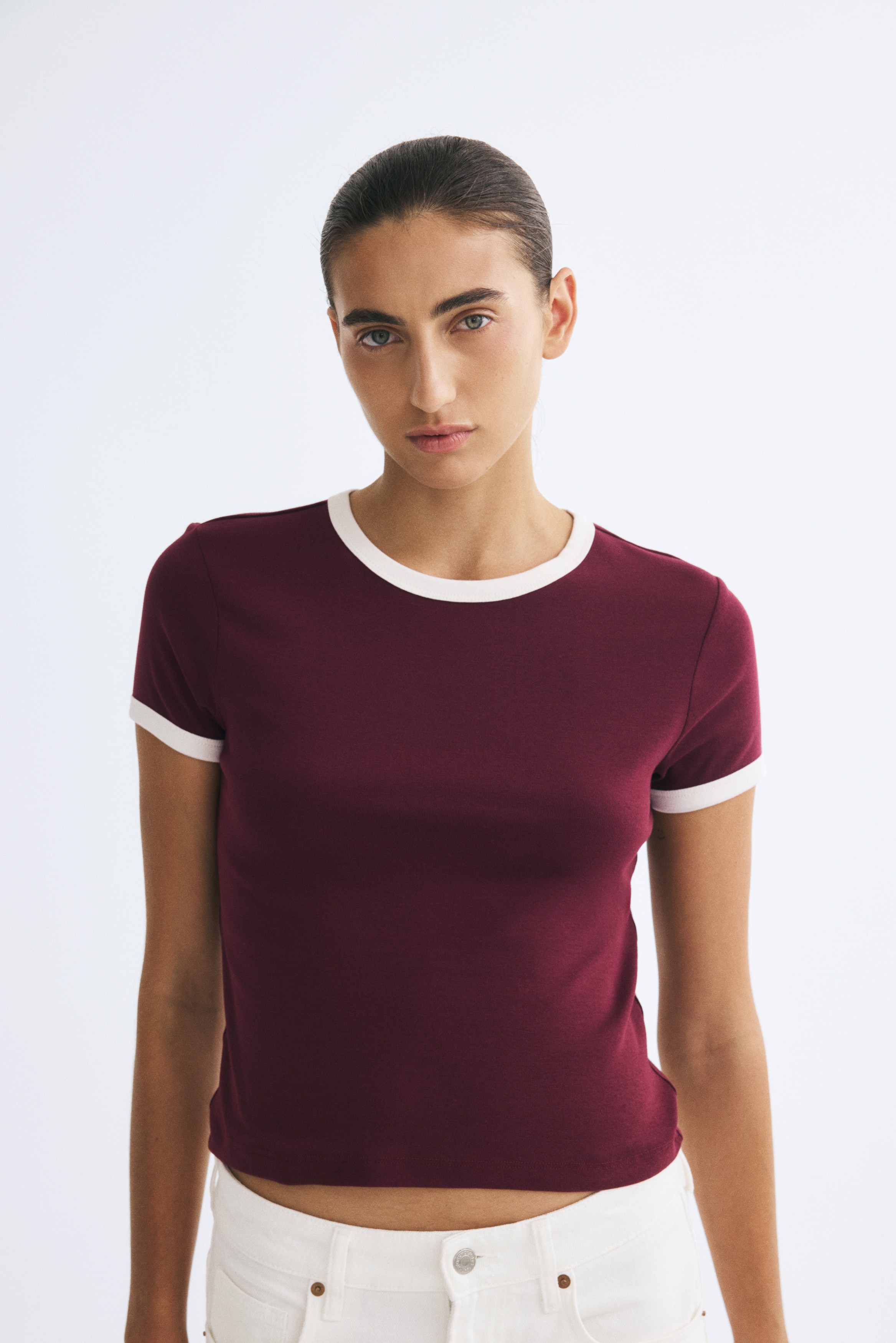 A burgundy fitted T-shirt is shown front-facing, featuring a round neckline and short sleeves with contrasting off-white trim on the cuffs, along with a straight-cut hem.