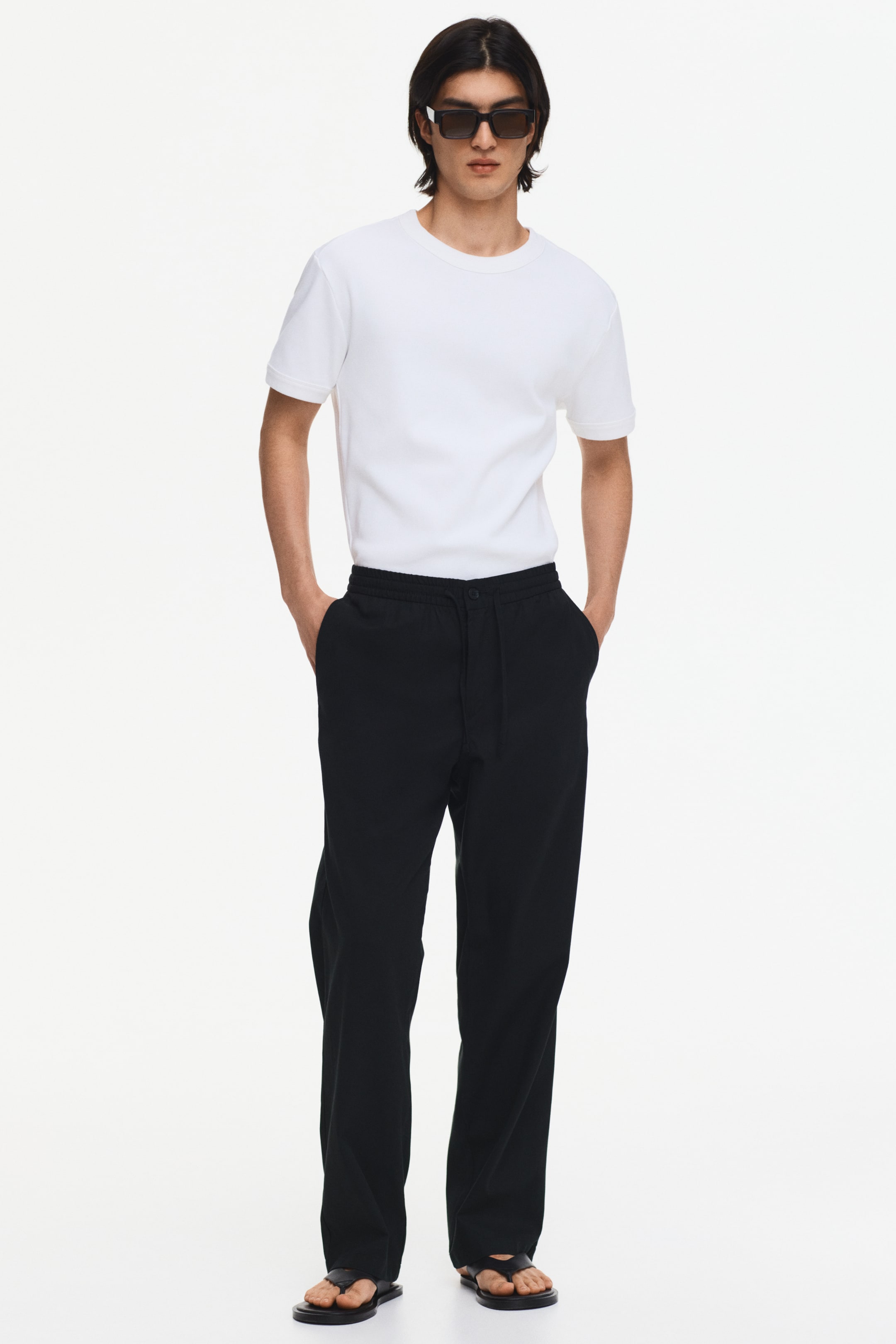 Agrandir l'image: Le t-shirt blanc présente un col rond classique, des manches courtes avec un ourlet net, et un tissu lisse et uni blanc. Sa coupe régulière est mise en valeur lorsqu'il est porté rentré dans un pantalon noir.