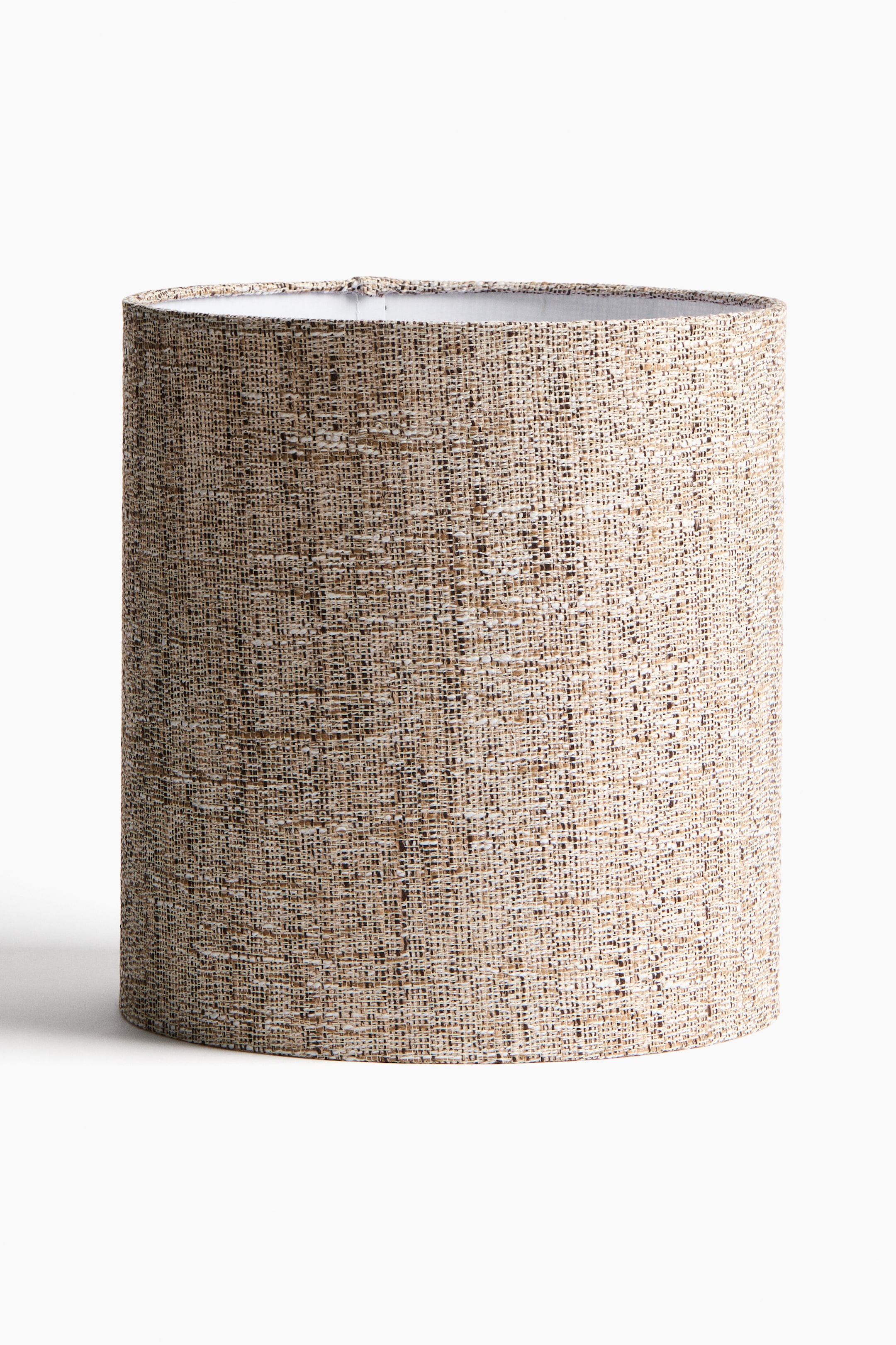 View larger image: A cylindrical lampshade is standing upright, angled slightly to the left. Its exterior is covered in a multi-tonal woven fabric featuring beige and brown threads, and a white interior lining is visible at the top opening.
