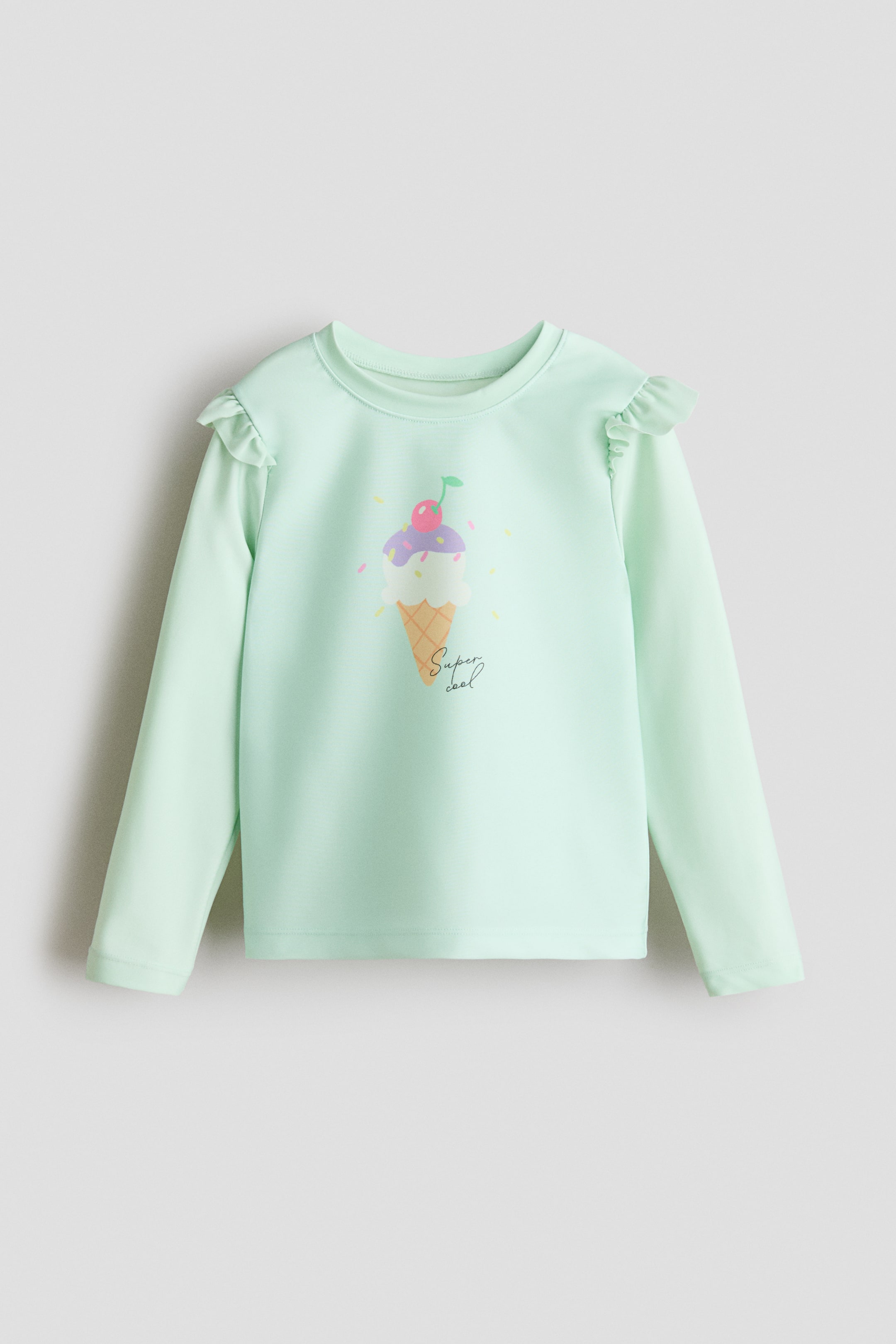 View larger image: A light green long-sleeved swim top is shown from the front, featuring a crew neck and frill-trimmed shoulders. An ice cream cone graphic with a cherry, sprinkles, and the text 'Super cool' appears on the front.
