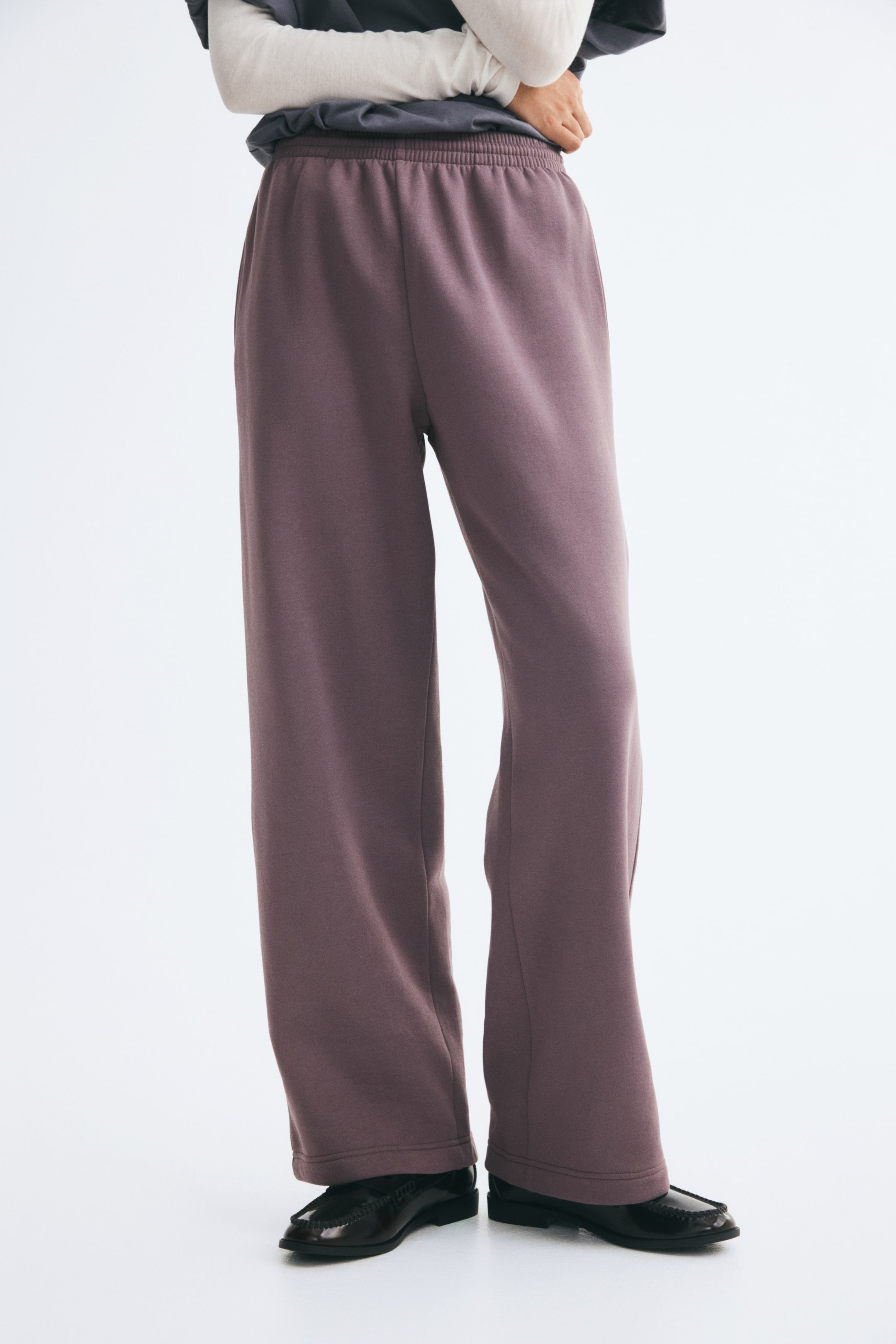 Größeres Bild anzeigen: Die von dem Model getragenen Trainingshosen aus roséfarbenem Sweatstoff verfügen über einen gesmokten elastischen Bund und gerade Beine. Das Model steht mit einer Hand in der Nähe der Taille und kombiniert die Hose mit übereinander getragenen Langarm-Oberteilen und schwarzen Loafern.