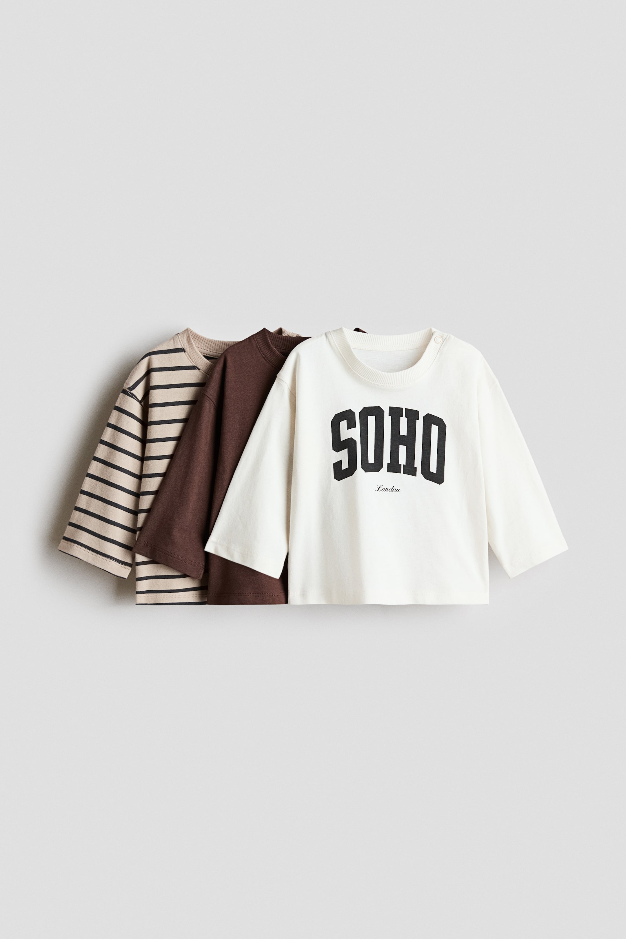 View larger image: Three long-sleeved t-shirts are layered from front to back, all featuring ribbed crew necks. The frontmost t-shirt is cream with a black 'SOHO London' graphic and two shoulder snap buttons, while a solid brown t-shirt and a beige t-shirt with black horizontal stripes can be seen behind it.