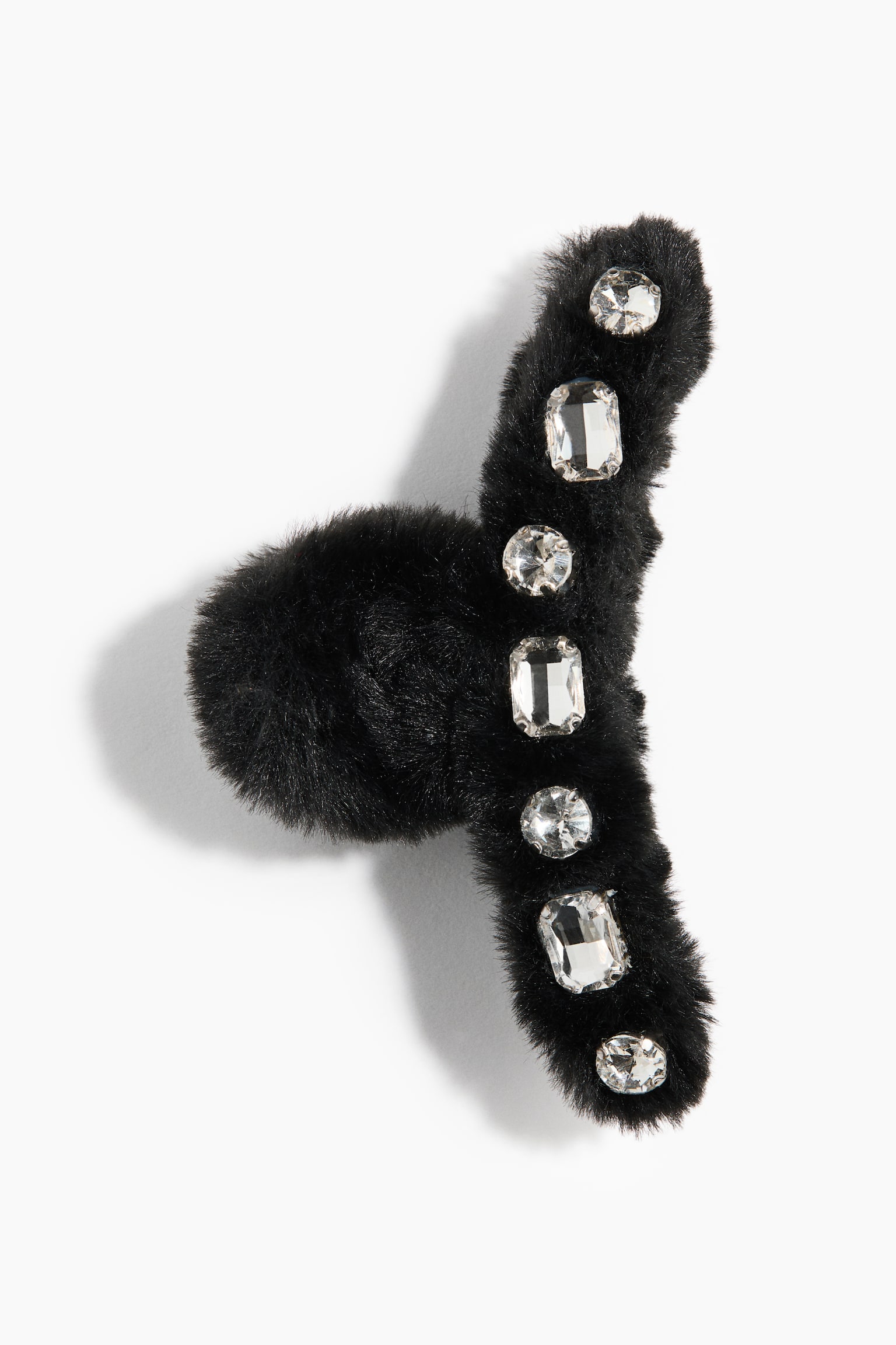 A black faux-fur claw hair clip displays a curved outer spine embellished with alternating round and emerald-cut clear crystals. The entire clip is covered in a soft, plush black fabric, highlighting its fuzzy texture and decorative gems.