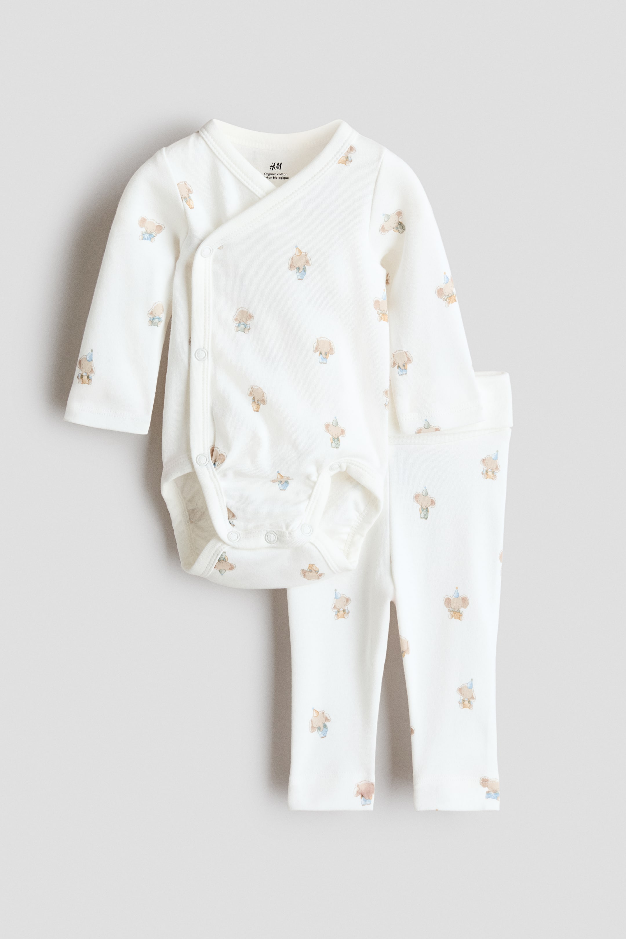View larger image: A two-piece white baby outfit includes a long-sleeved wrap-over bodysuit and matching long pants, both showcasing a repeating pattern of small, light-coloured animal figures. The bodysuit is designed with snap closures along the side and at the crotch, while the pants feature an elastic waistband.