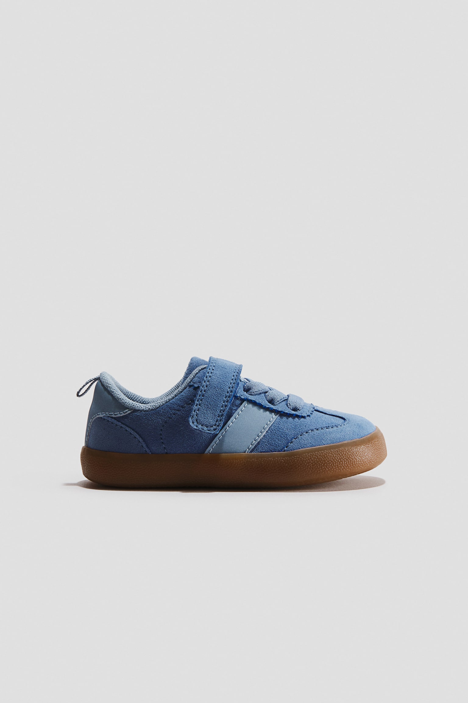 Barefoot trainers are facing right in a side profile view, featuring a blue suede upper with a lighter blue diagonal side stripe, elastic laces, and a single hook-and-loop strap closure. The shoe displays a wide, rounded toe box, a heel pull tab, and a textured brown gum sole.