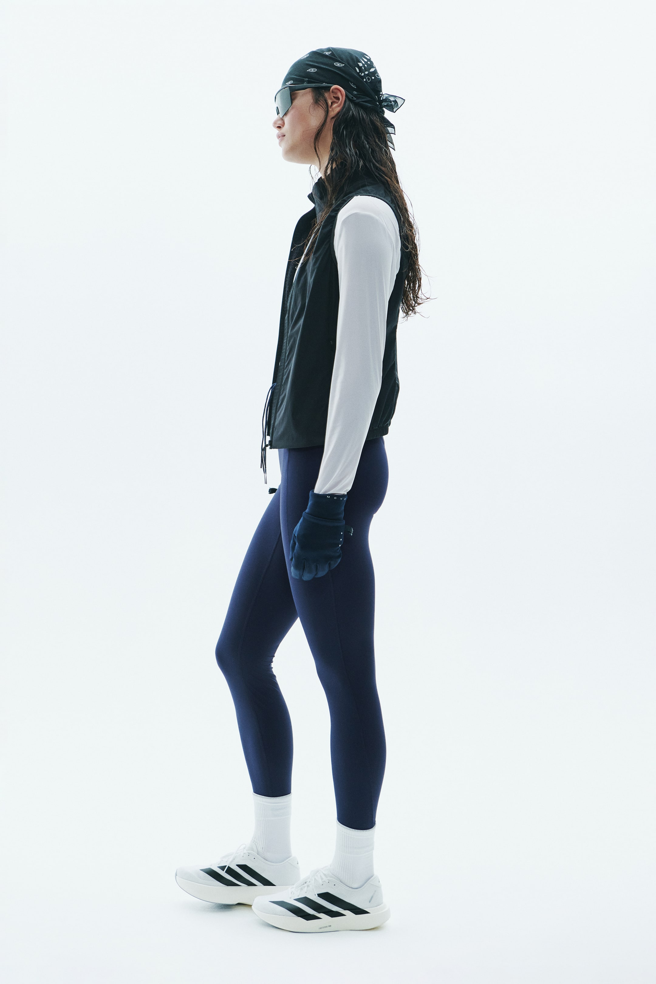 Voir une image plus grande: Un gilet noir sans manches à fermeture éclair, doté d’un col montant et d’un ourlet à cordon de serrage, est porté par le mannequin. Le gilet est superposé à une chemise blanche à manches longues et agencé avec des leggings bleu marine et des chaussures de sport blanches.