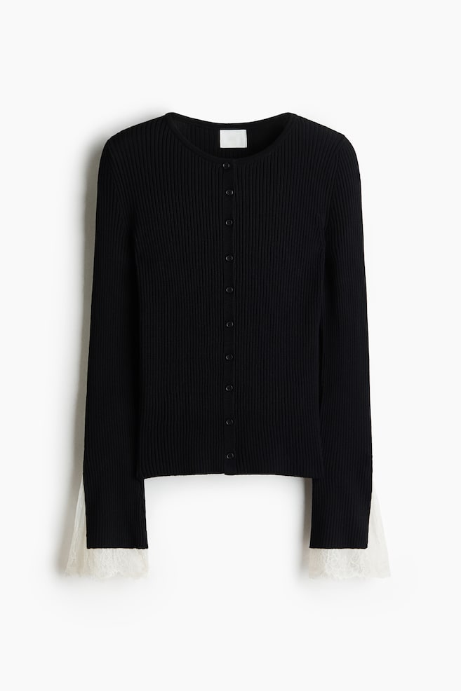 Eine schwarze Strickjacke mit Rippenstruktur wird von vorne gezeigt und verfügt über einen runden Ausschnitt sowie einen Knopfverschluss mit kleinen, farblich abgestimmten Knöpfen. Die langen Ärmel enden in ausgestellten Manschetten mit Schlitz, akzentuiert durch kontrastierende weiße, gewellte Spitzenbesätze. - 1