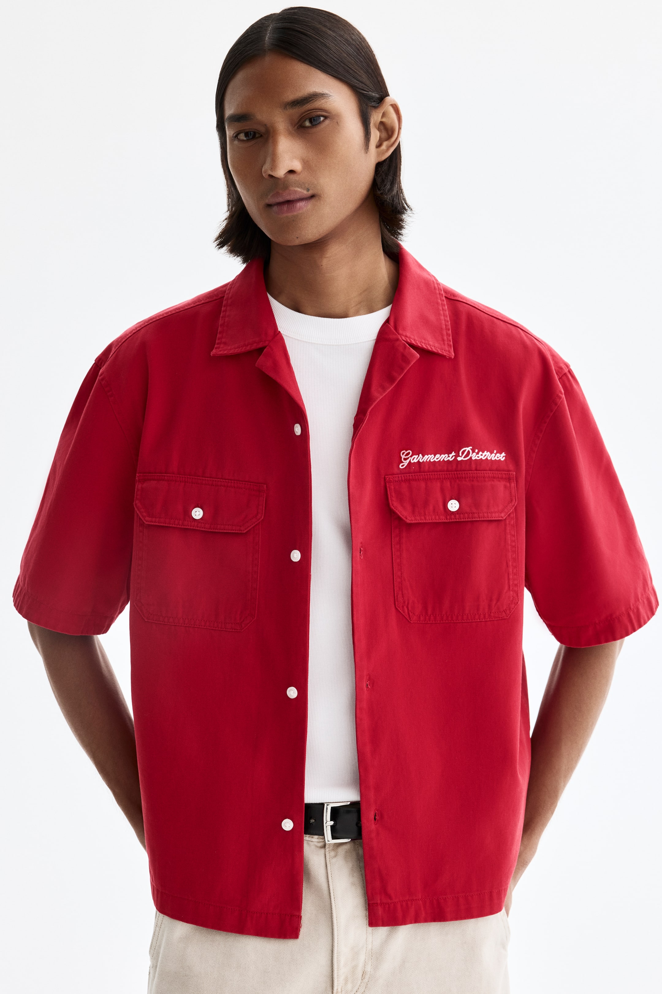 View larger image: A red short-sleeved, button-front shirt faces forward, featuring a Cuban collar and two chest flap pockets with white buttons. White embroidery reading "Garment District" details the left chest, and the shirt is partially unbuttoned, revealing a white ribbed T-shirt underneath.