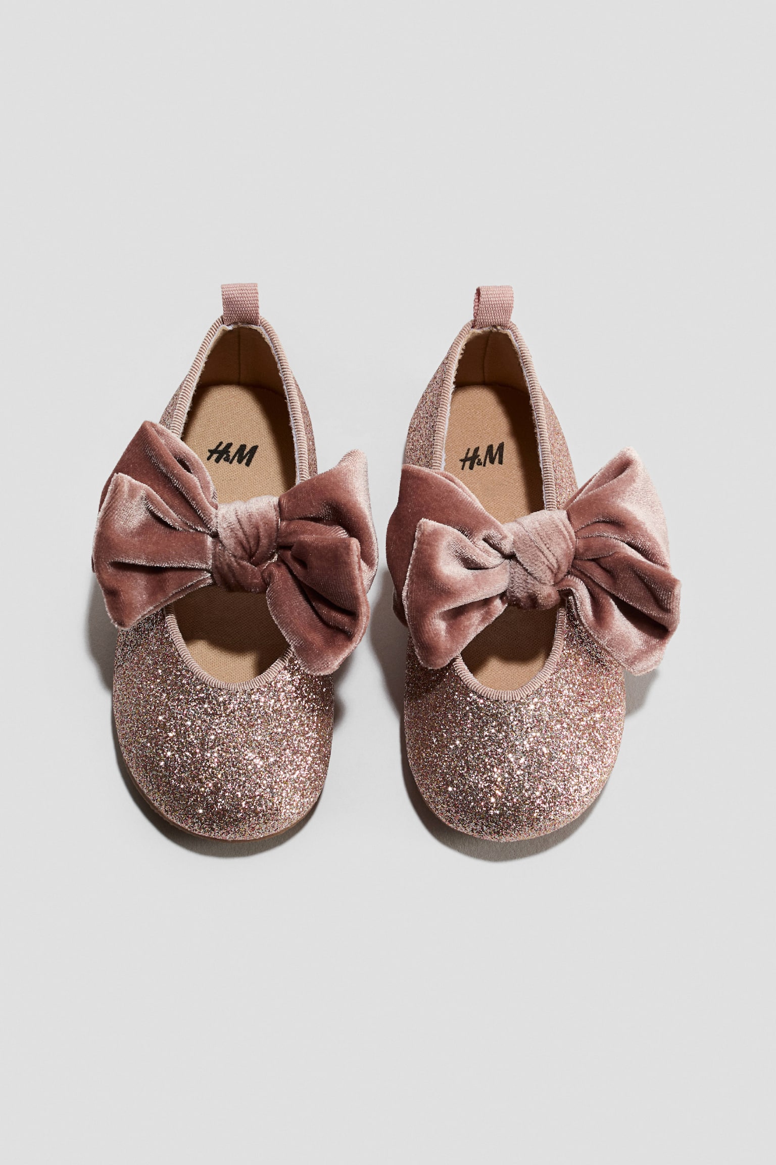 A pair of pink glittery ballet flats is shown from a top-down view, featuring large, plush dusty pink velour bows on the straps across the instep. The shoes are designed with small fabric pull tabs at the heels and beige insoles.