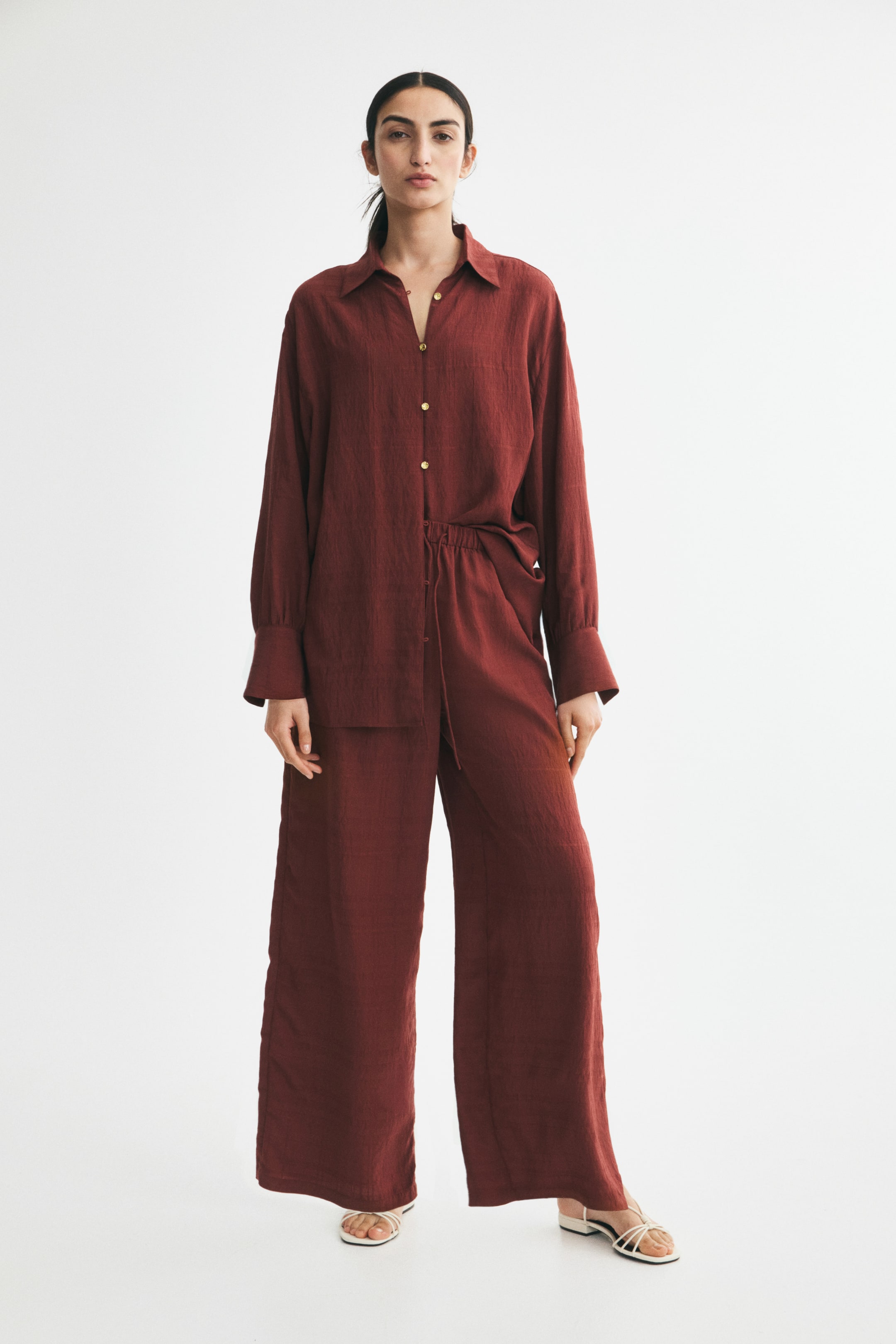 View larger image: Wide-leg trousers in a textured, rust-coloured weave are worn by the model, featuring a relaxed fit and an elasticated waistband with a hanging drawstring. The trousers are styled with a matching long-sleeved button-up shirt and white strappy sandals.