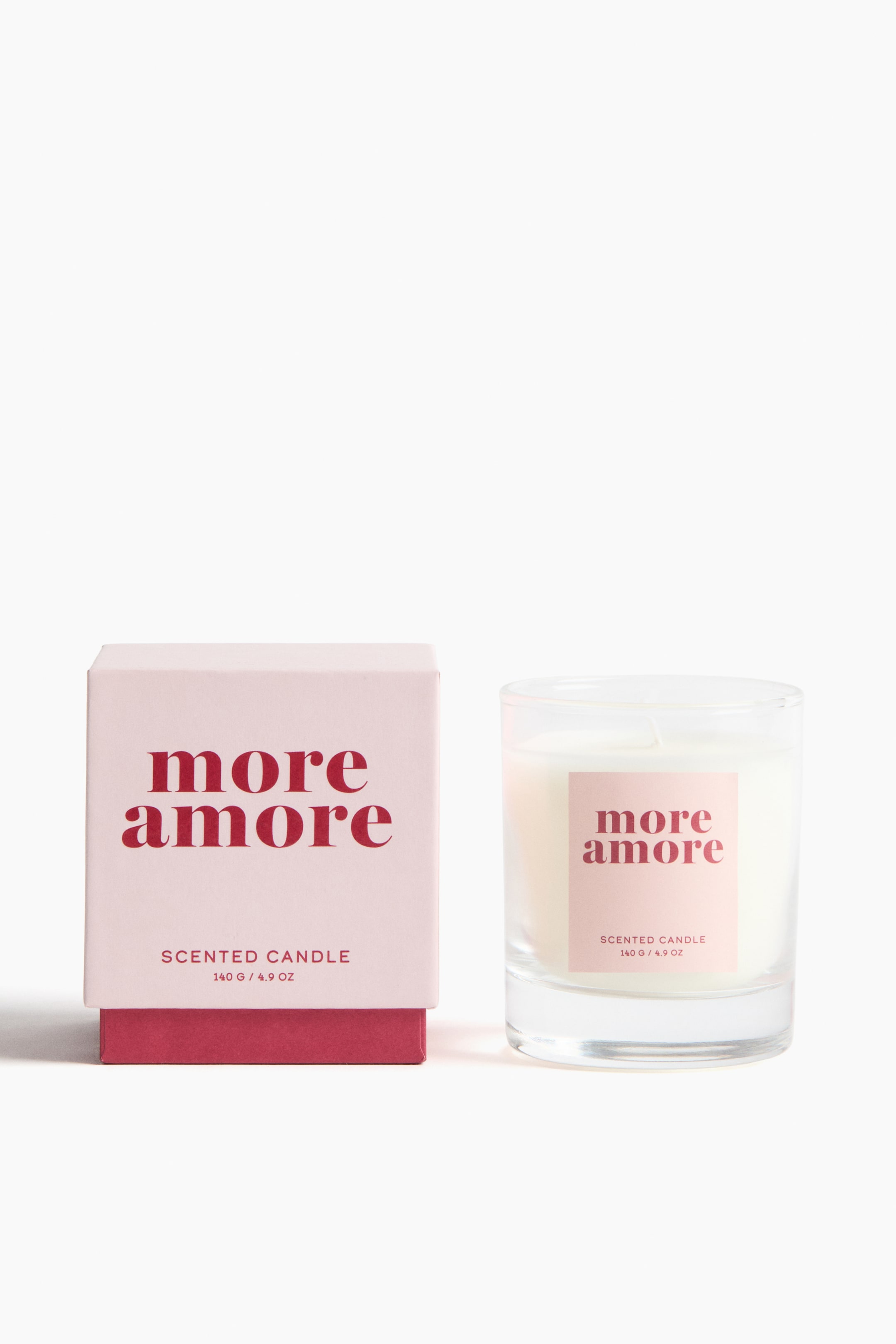 View larger image: A scented candle in a clear glass jar contains off-white wax and displays a light pink label on its front. It is presented alongside its matching light pink product box, which features a dark pink base and stands open to the left of the candle. Both the candle and box labels read 'more amore' and 'scented candle 140 G / 4.9 OZ' in dark pink text.