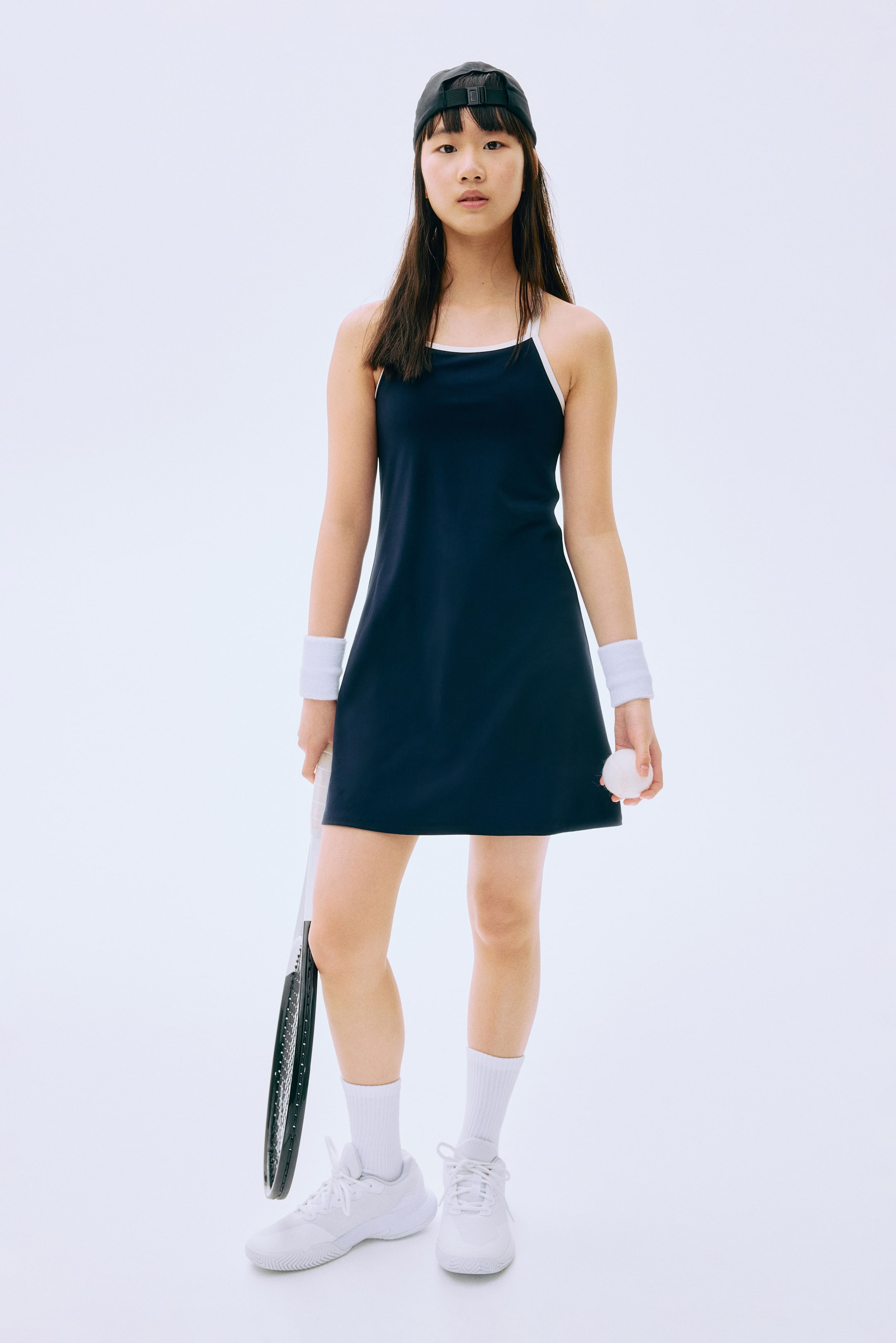 Größeres Bild ansehen: Ein marineblaues Tenniskleid wird von vorne präsentiert, mit einem Rundhalsausschnitt und dünnen Trägern mit weißem Besatz. Ein weißer Sport-BH ist sichtbar darunter geschichtet, und das Kleid ist in einer A-Linien-Silhouette geschnitten, die über den Knien endet.