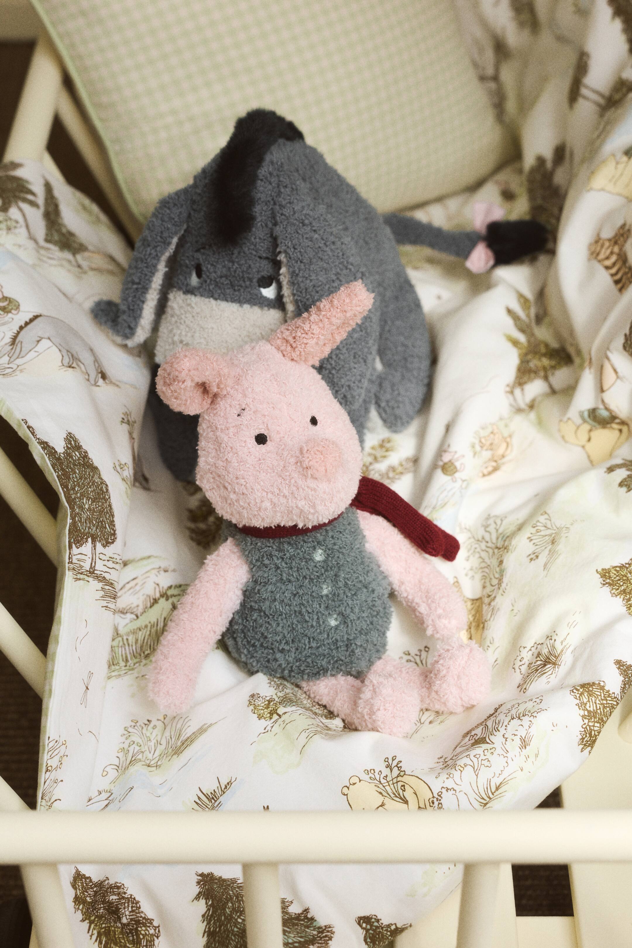 拡大画像を表示: ピンクのぬいぐるみの豚のおもちゃが、ボタン付きのダークグレーのベストと赤いニットのスカーフを身に着けて直立しています。その後ろには、黒いたてがみとピンクのリボンが付いたしっぽを持つグレーのぬいぐるみのロバのおもちゃが横たわっています。