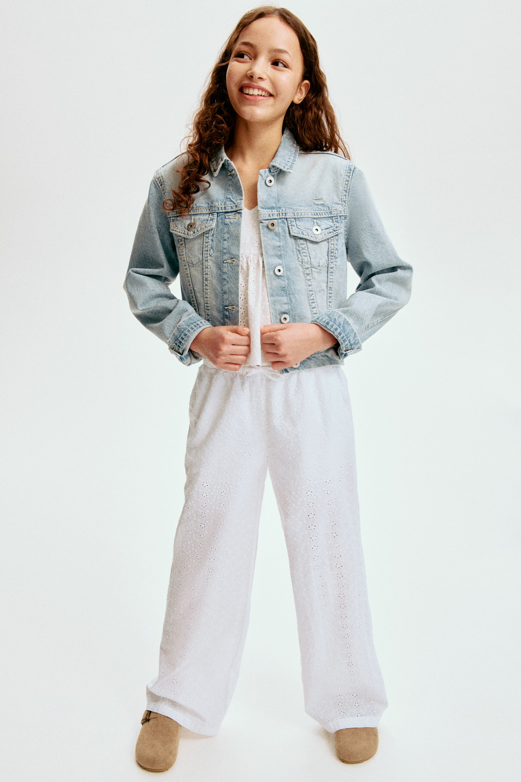 View larger image: A light blue denim jacket featuring a pointed collar, metal button closures down the front, two chest flap pockets, two side pockets, a faded wash, a banded hem, and long sleeves with buttoned cuffs, worn open over a white top.