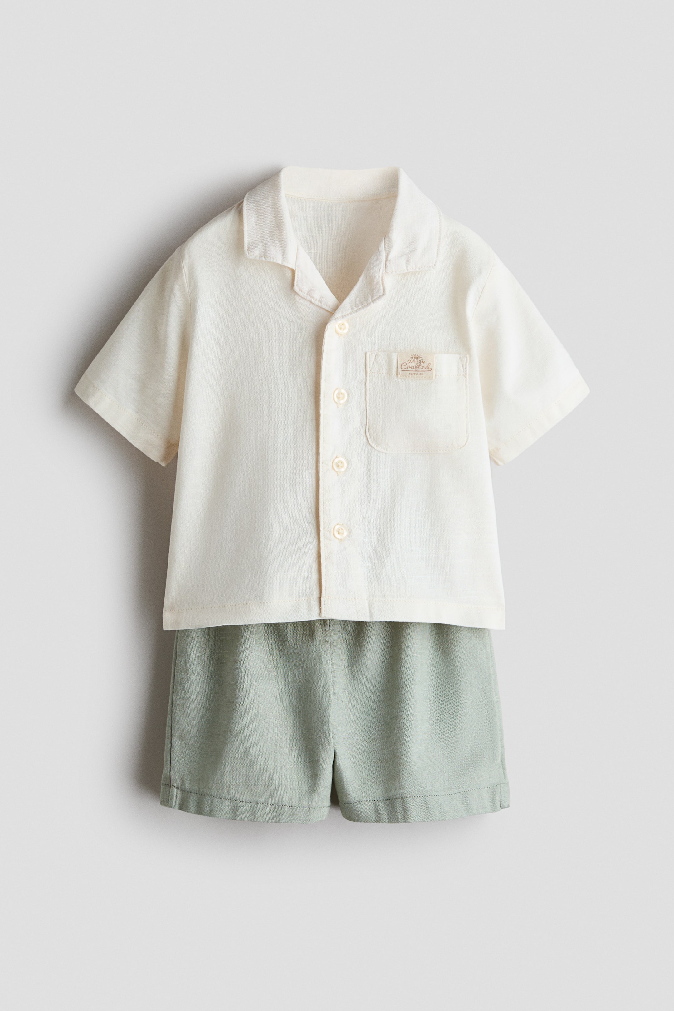View larger image: A two-piece set ditunjukkan dari hadapan, terdiri daripada kemeja lengan pendek berwarna putih pudar dengan kolar camp, butang di bahagian depan, dan poket tampal di dada dengan label sulaman kecil. Kemeja ini dilapiskan di atas seluar pendek berwarna hijau muda.