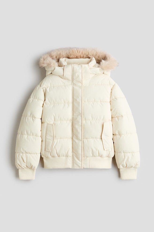 A cream-coloured puffer jacket is shown from the front, featuring horizontal quilting and a hood trimmed with light brown faux fur. The jacket includes a concealed central zip with a storm flap, two slanted side pockets, and ribbed knit cuffs and hem. - 1