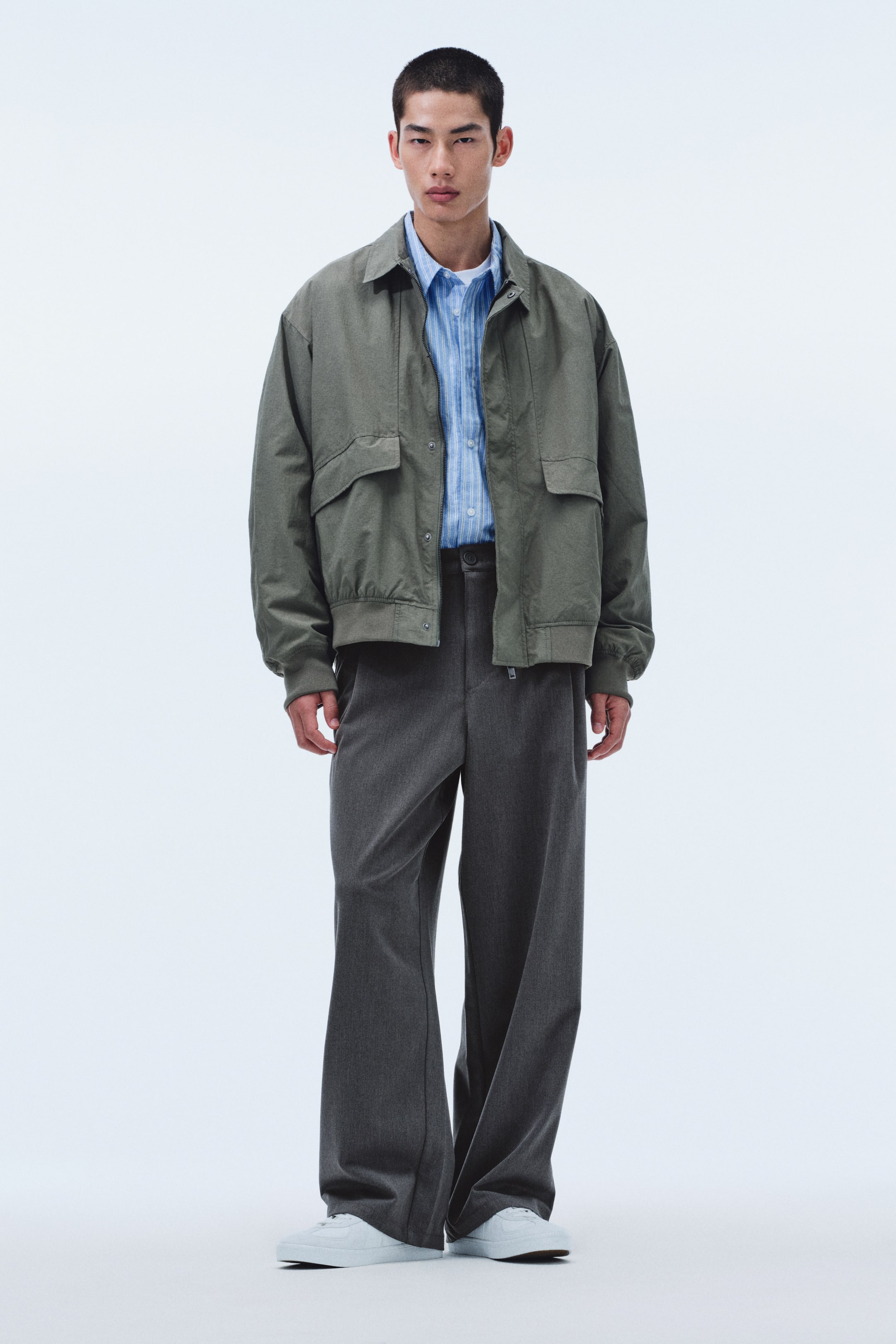View larger image: An olive-green bomber jacket features a pointed collar, two chest flap pockets, and elasticated hem and cuffs, worn open over a light-blue striped button-down shirt. Dark-gray wide-leg trousers with a visible drawstring waist complement the jacket, and white sneakers complete the ensemble.
