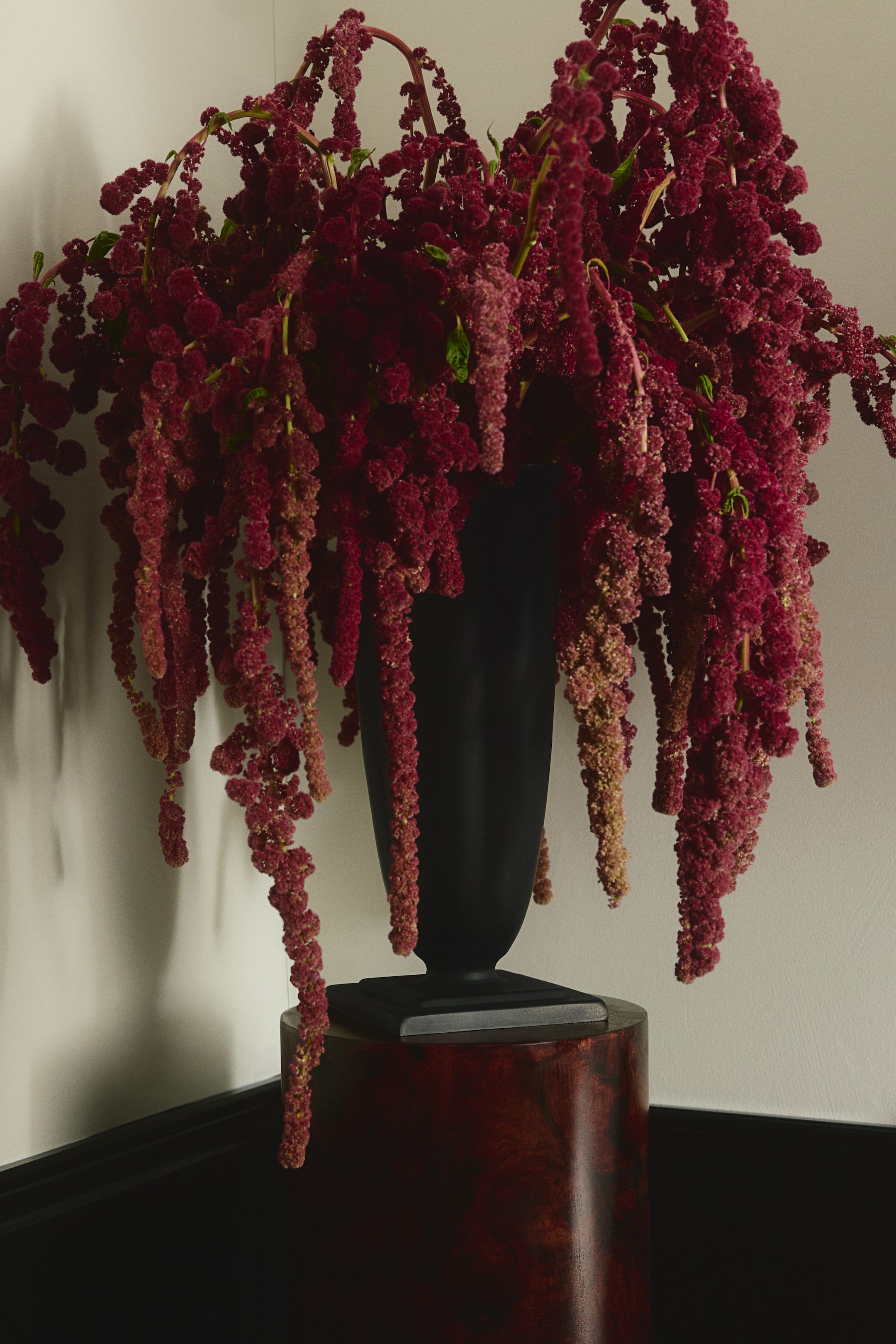 View larger image: A large black metal vase facing forward rests on a polished cylindrical pedestal, featuring a tapered body, a fluted rim, and a sculptural, square-stepped foot. Cascading burgundy amaranth flowers spill from the vase and drape over its sides.