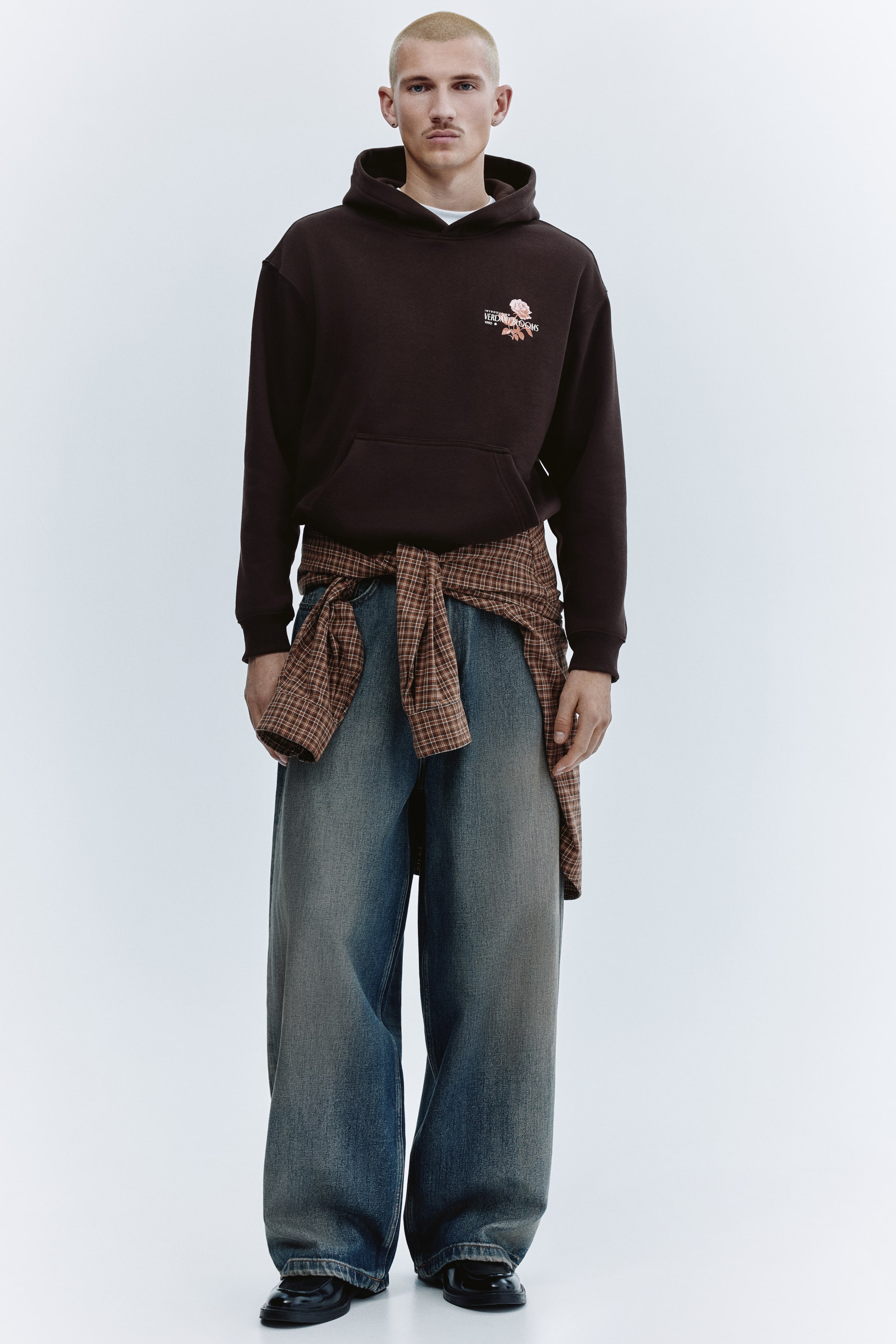 이미지 확대 보기: 다크 브라운 컬러의 오버사이즈 후디로, 왼쪽 가슴에 텍스트와 장미의 작은 그래픽 프린트가 있으며 캥거루 포켓이 특징입니다. 후드와 드로스트링, 시보리 처리된 소맷단 및 밑단으로 디자인되었고, 그 아래로 화이트 티셔츠가 보입니다.