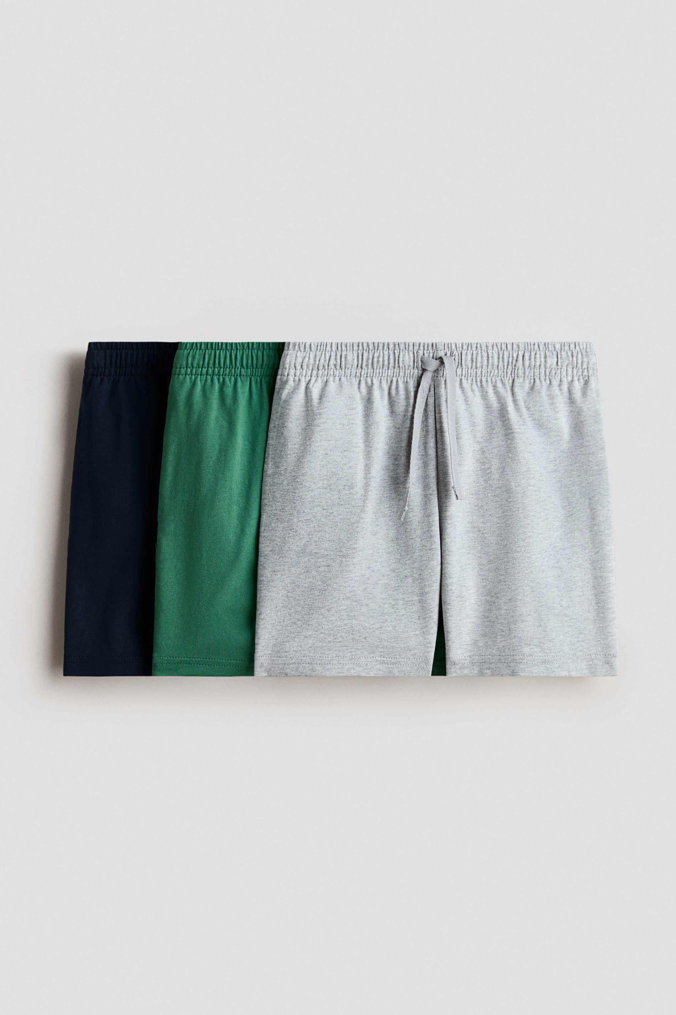 View larger image: Three pairs of shorts in soft cotton jersey are showcased, each featuring an elastic, drawstring waist. The light gray marl shorts are fully visible from the front, highlighting the drawstring, while the partially visible green and navy blue shorts are positioned alongside.