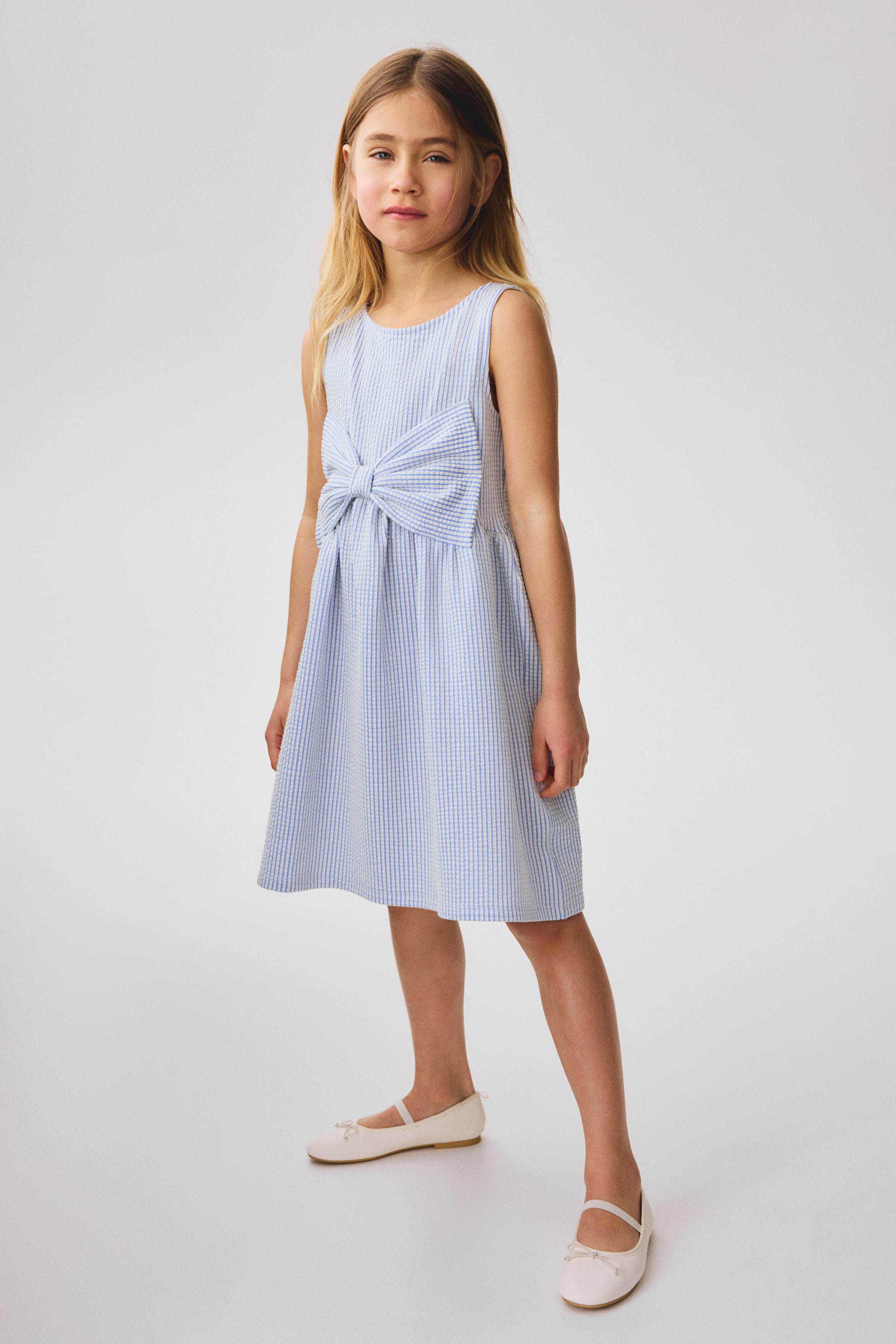 Näytä suurempi kuva: Hihaton mekko seersucker-tyylisestä jersey-kankaasta, jossa on pyöreä pääntie ja edessä suuri, kiinteä rusetti, joka on sijoitettu vyötäröllä olevan poimutetun sauman yläpuolelle, josta levenee helmaan.