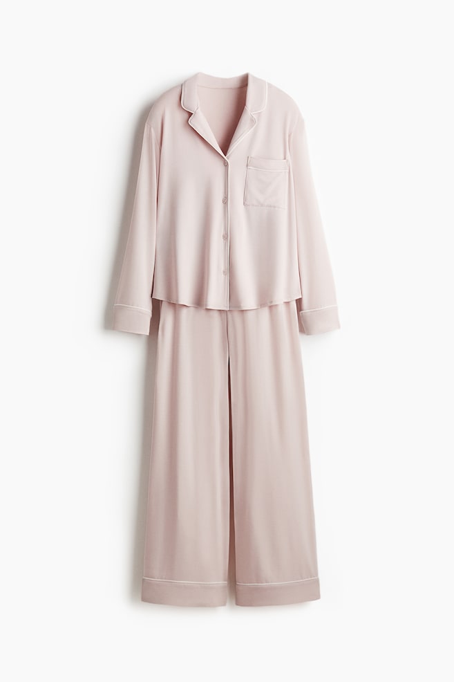 A light pink pyjama set is being displayed, featuring a long-sleeved shirt and matching wide-leg bottoms. The shirt, facing FRONT, is designed with white-piped notch lapels, buttons down the front, and a visible left chest pocket. The bottoms are showing a smooth waistband and wide, straight legs with white piping at the hems. - 1