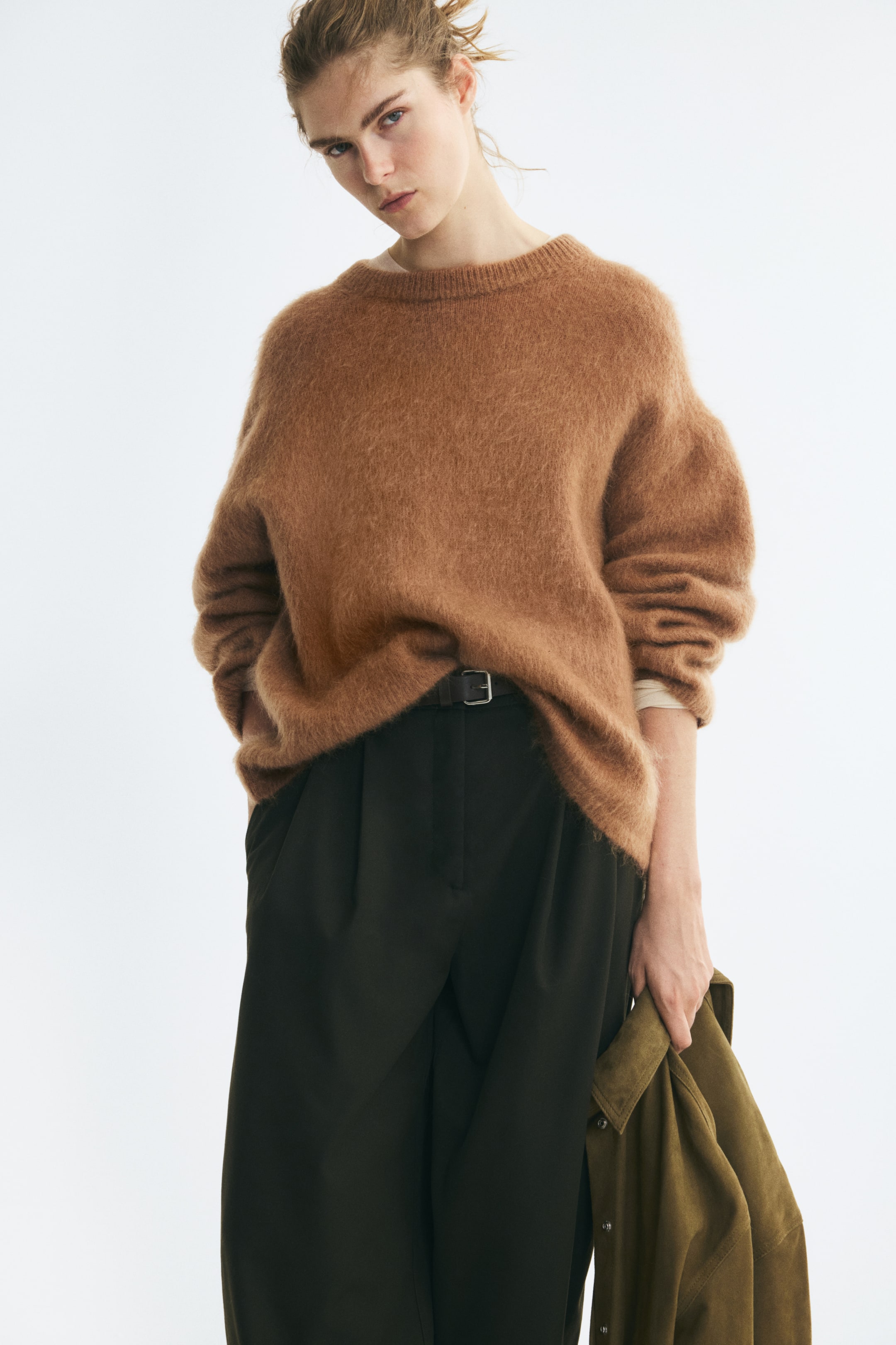 Vis større bilde: En oversized, brun, mykstrikket genser med senkede skuldre og ribbestrikket hals er brukt av modellen. Genseren er stylet delvis stukket ned i mørke bukser i livet, mens modellen holder en grønn jakke.