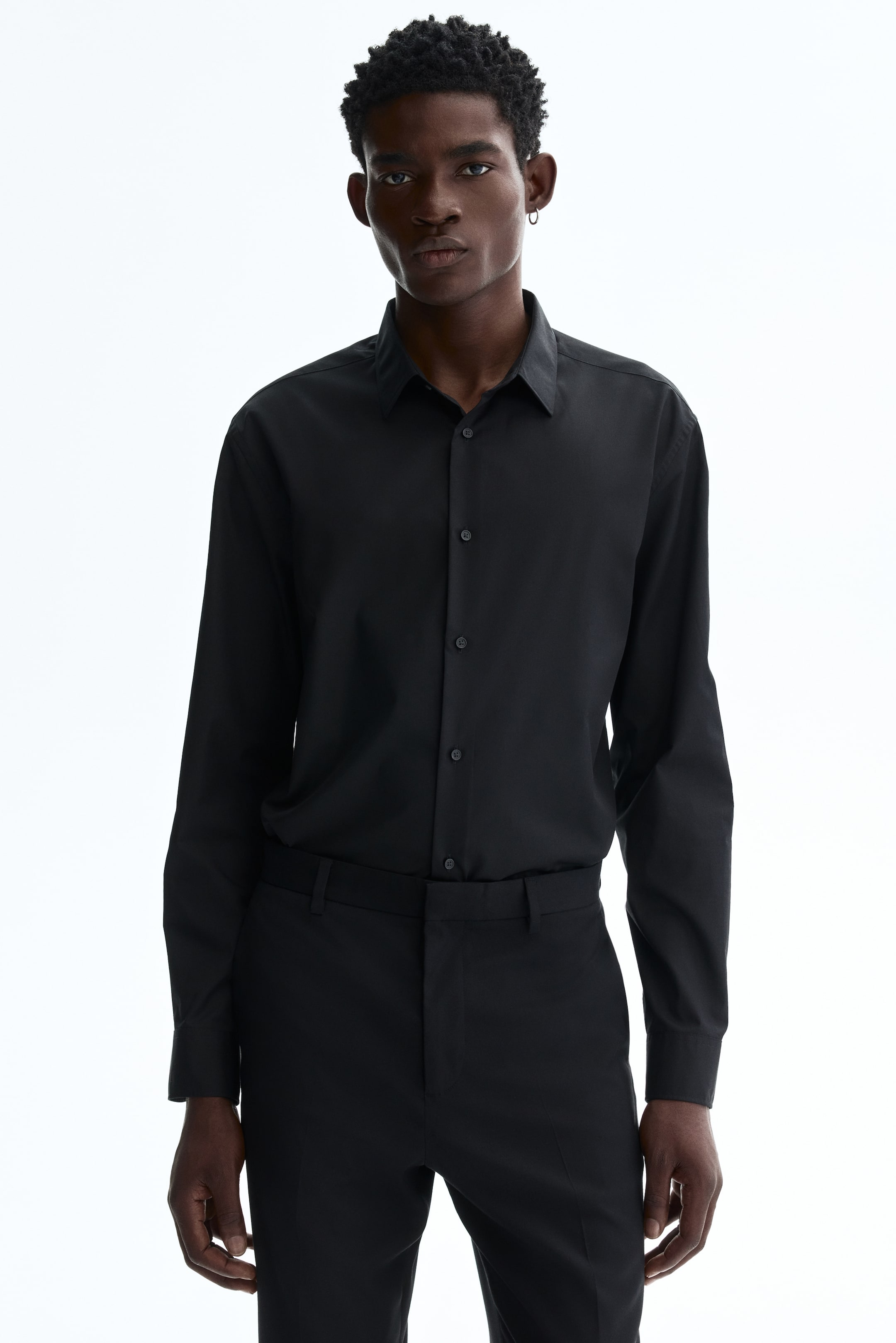View larger image: A black button-down shirt is showcased from the front, featuring a classic pointed collar and a button-front placket with visible black buttons. Its long sleeves end in buttoned cuffs, and the shirt fits smoothly, tucked into black trousers.