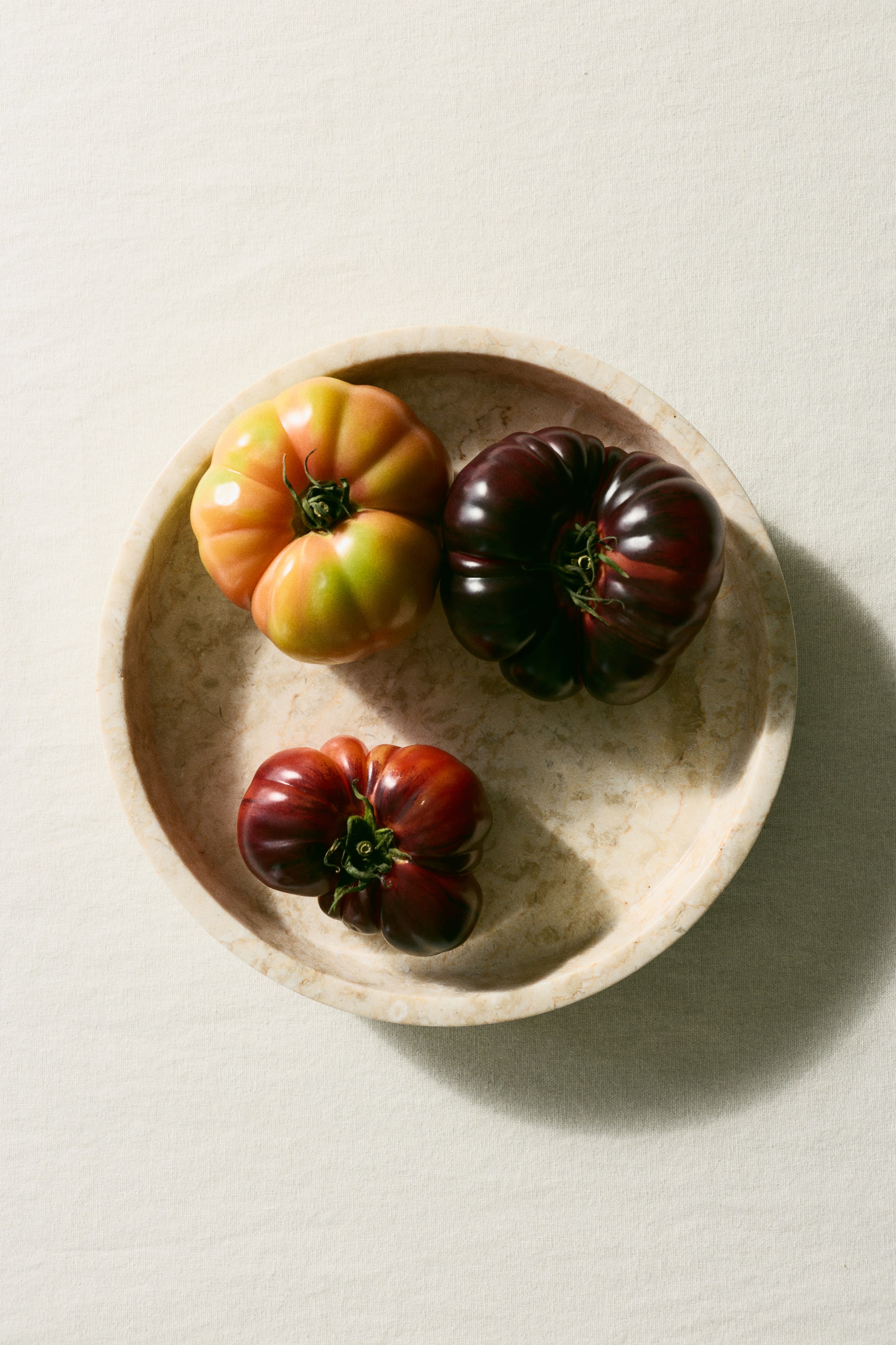 View larger image: Three irregularly shaped heirloom tomatoes are resting in a shallow, light-coloured stone bowl, viewed from above. One tomato is mottled orange and green, another is deep purplish-red, and the third is reddish-purple, all exhibiting deep ribbing.
