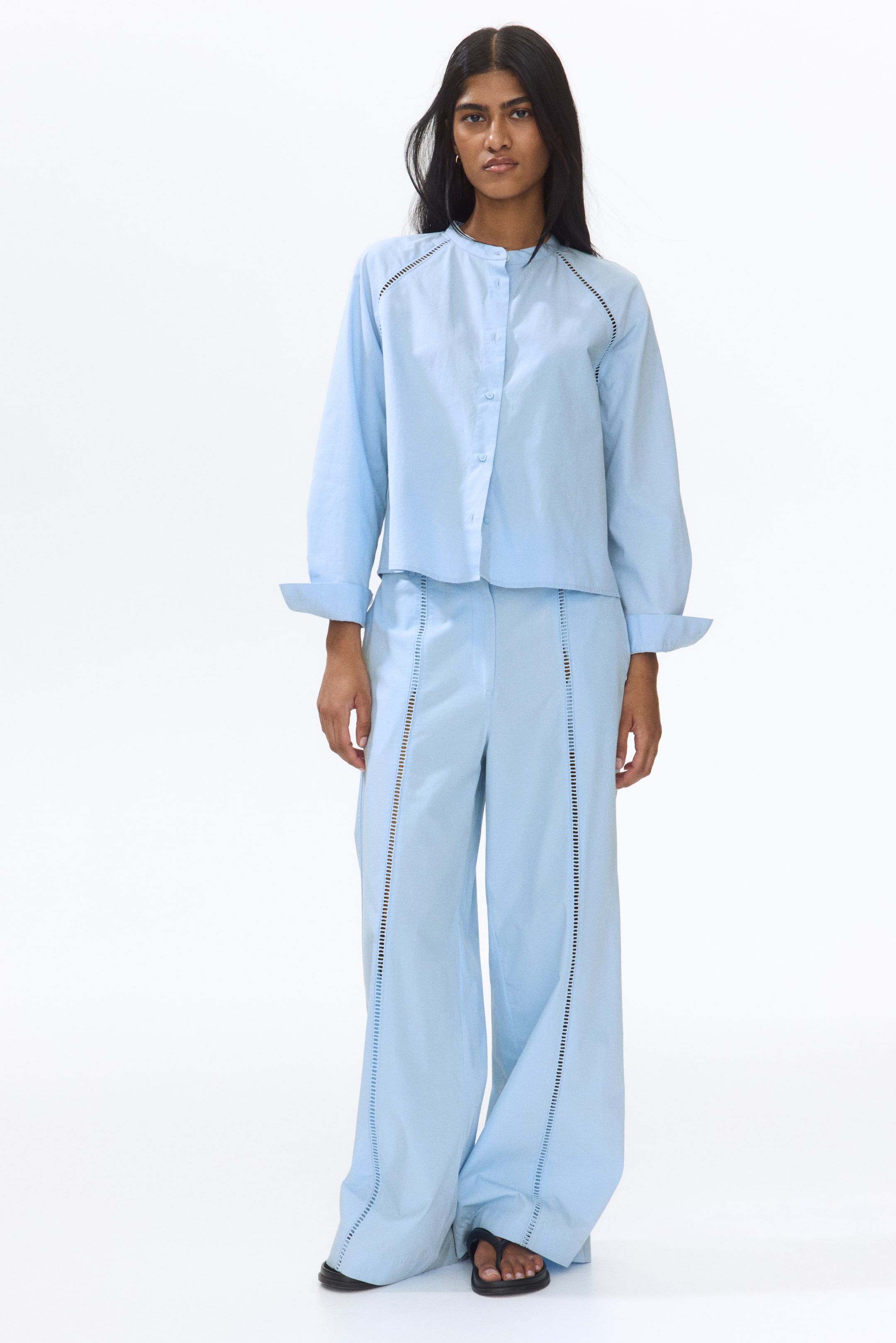 View larger image: The model is wearing a light blue, loose-fit blouse with ladder-lace insets along the raglan seams. It features a band collar and buttons down the front, with long sleeves that have cuffs rolled up. The blouse is styled with matching light blue trousers.