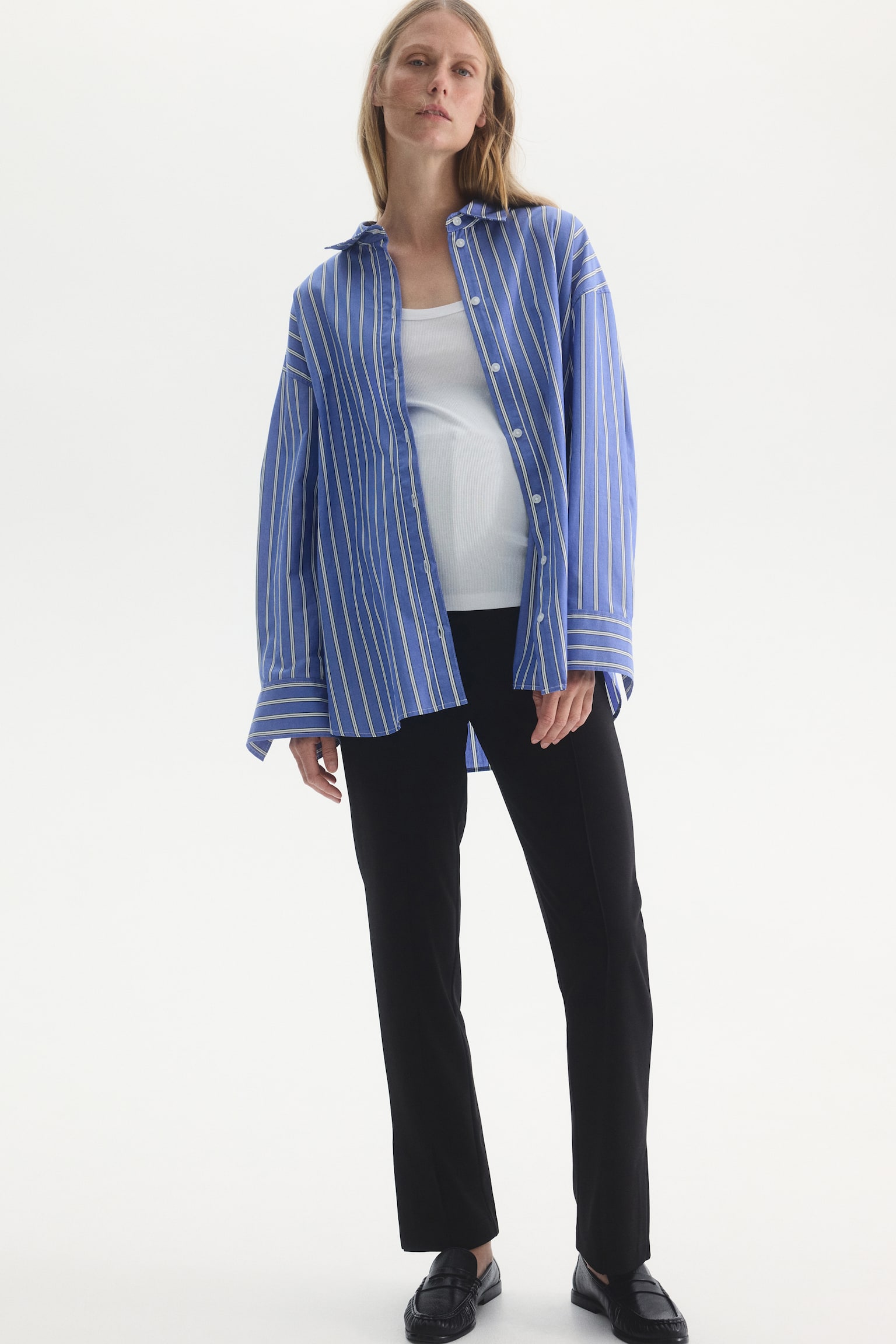 The model is wearing black cigarette trousers, featuring a slim, straight-leg silhouette with front creases. The trousers are styled with a white top under an open blue and white striped button-up shirt, and black loafers. - 1