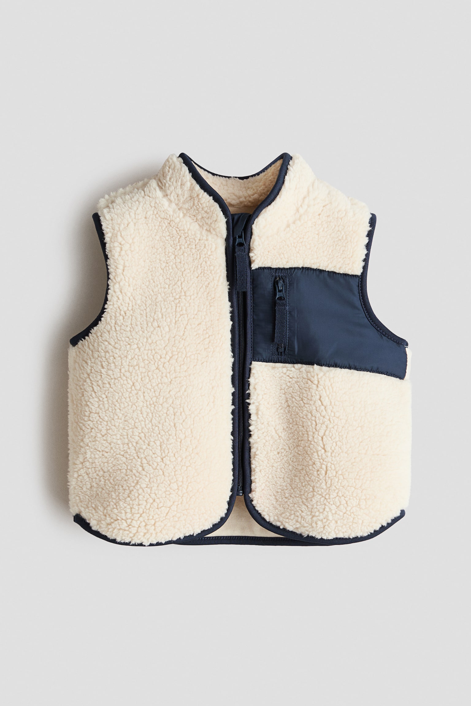 Un chaleco de peluche color crema se muestra de frente, con un cuello alto y un cierre de cremallera azul marino en la parte delantera con protector de barbilla. Está diseñado con un bolsillo con cremallera azul marino en tela tejida en la parte superior derecha, y los ribetes de jersey azul marino adornan el cuello, las sisas y el dobladillo.