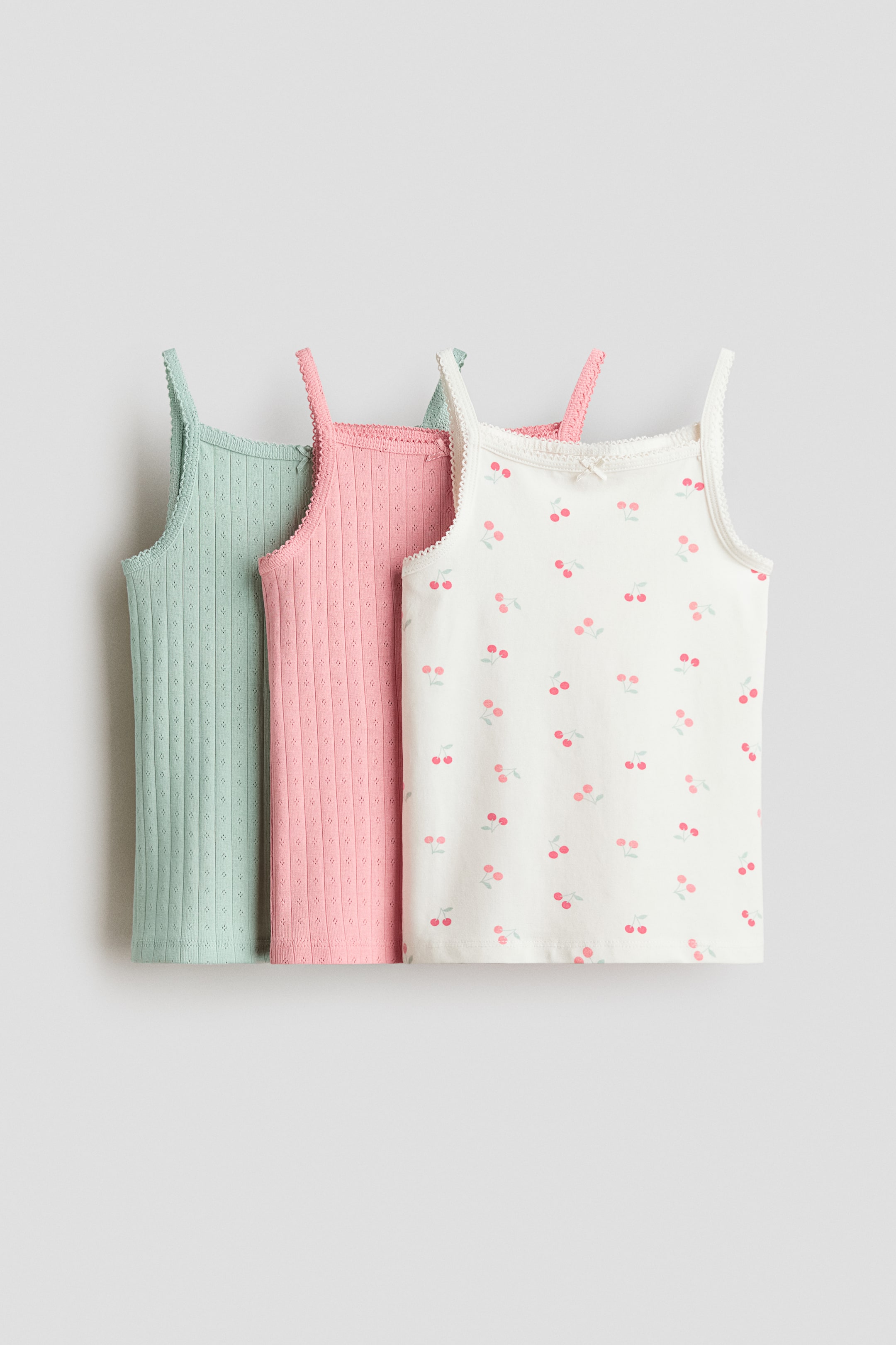 View larger image: Three FRONT tops in soft cotton jersey feature square necklines with picot trims and narrow shoulder straps. One white top displays a cherry pattern with a decorative satin bow at the neckline, while the other two, in light green and pink, present a ribbed texture with small perforations.