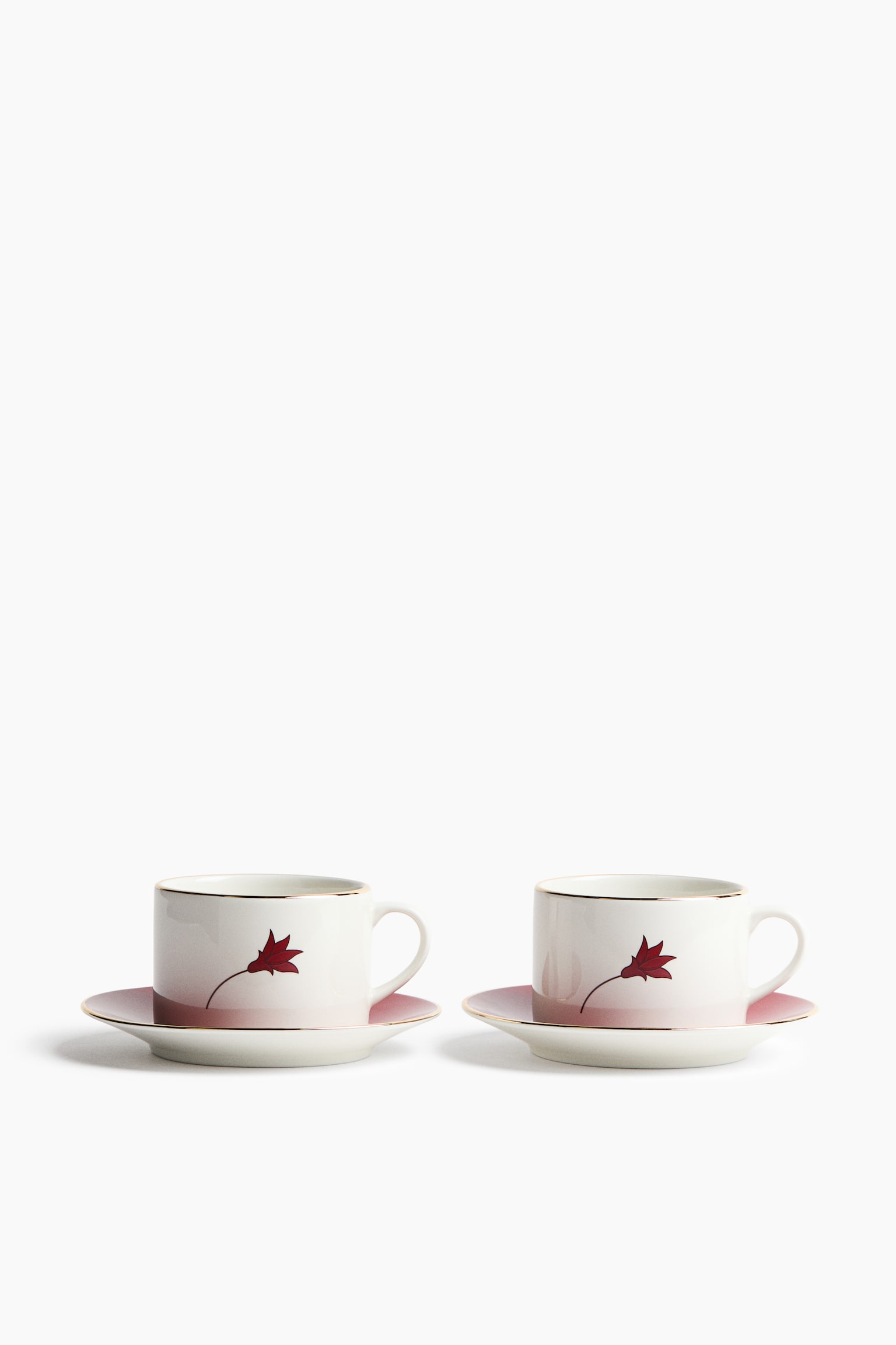 A set of two white ceramic teacups with matching saucers is displayed from a front angle, sitting side-by-side. Each cup and saucer features a pink ombrÃ© gradient fading upwards, gold-gilded rims, and a single stylised red flower with a delicate stem printed on the side of the cup.