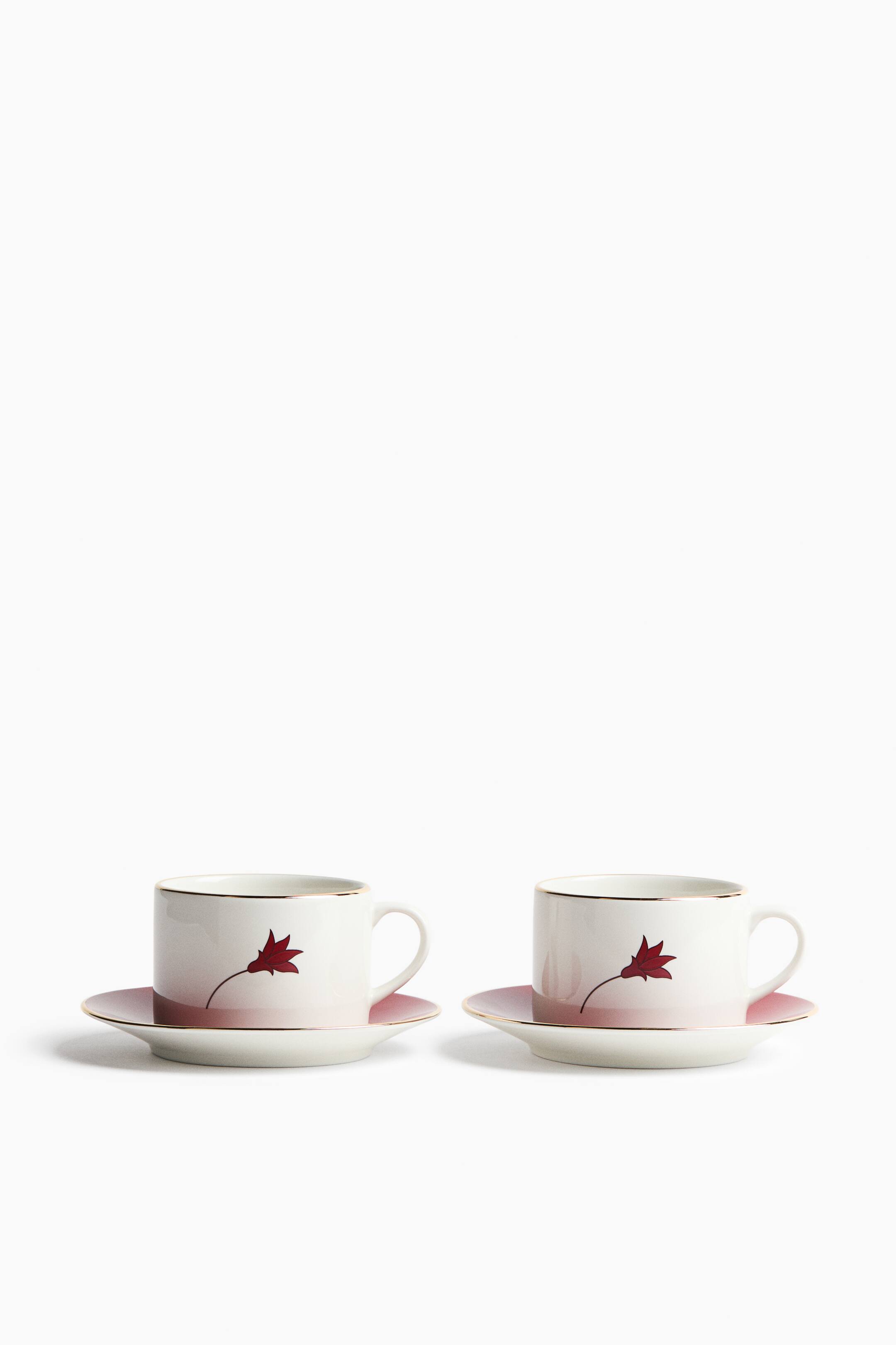 View larger image: A set of two white ceramic teacups with matching saucers is displayed from a front angle, sitting side by side. Each cup and saucer features a pink ombré gradient fading upwards, gold-gilded rims, and a single stylised red flower with a delicate stem printed on the side of the cup.