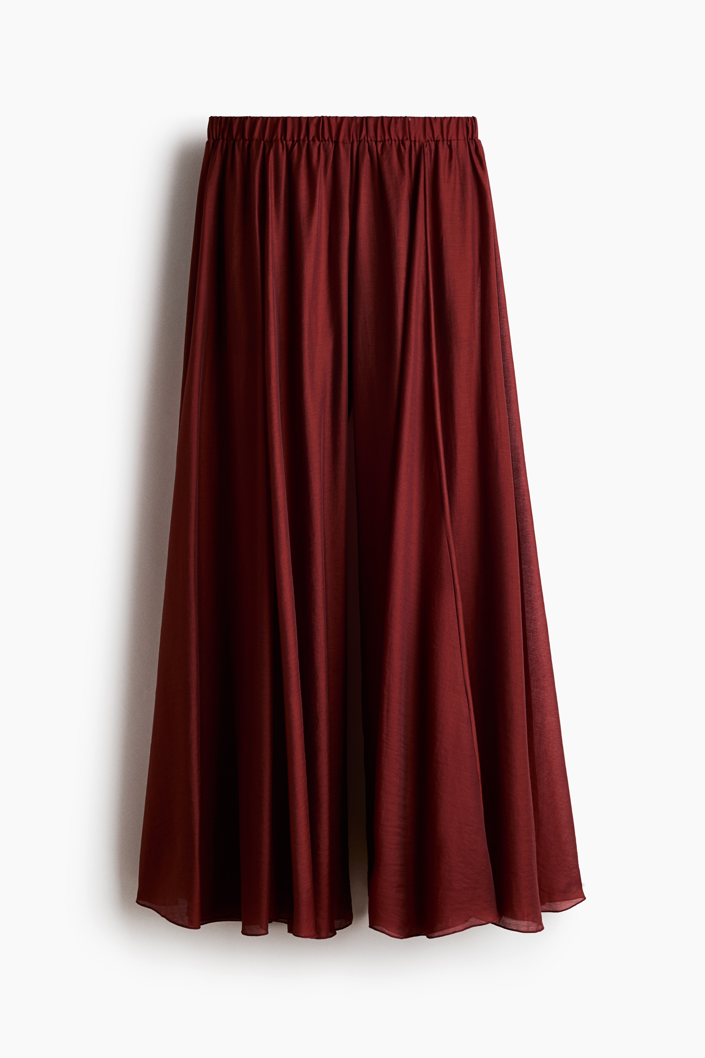 Oversized, wide-leg pants in a sheer red fabric are being worn by the model, who is standing with their back to the camera. The pants feature a flared silhouette with a shorter lining visible underneath and are styled with a matching ruffled camisole top.