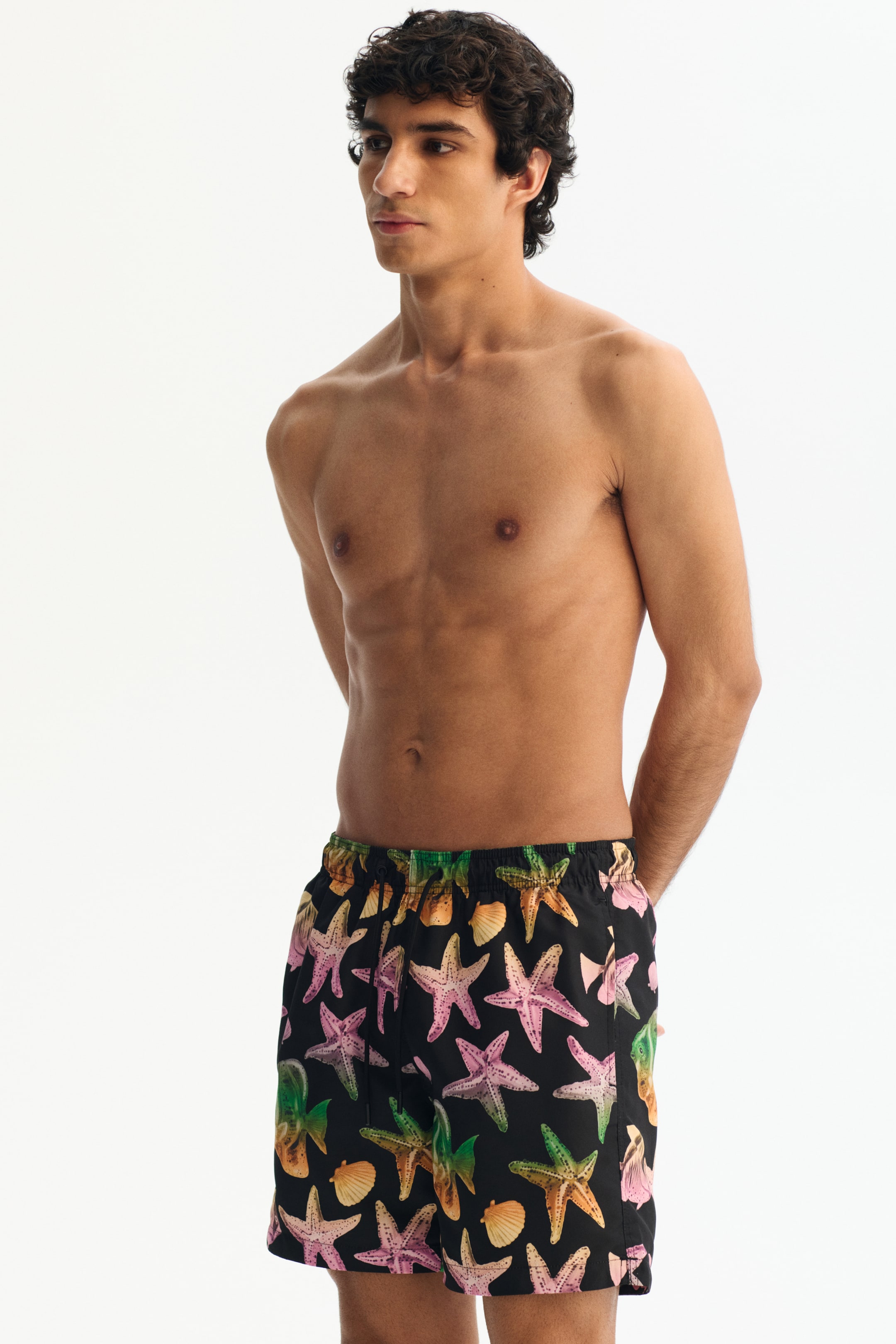 Agrandir l'image: Le mannequin porte un short de bain. Le short noir affiche un imprimé intégral de coquillages et d'étoiles de mer colorés, et est conçu avec une taille élastique et un cordon de serrage visible.