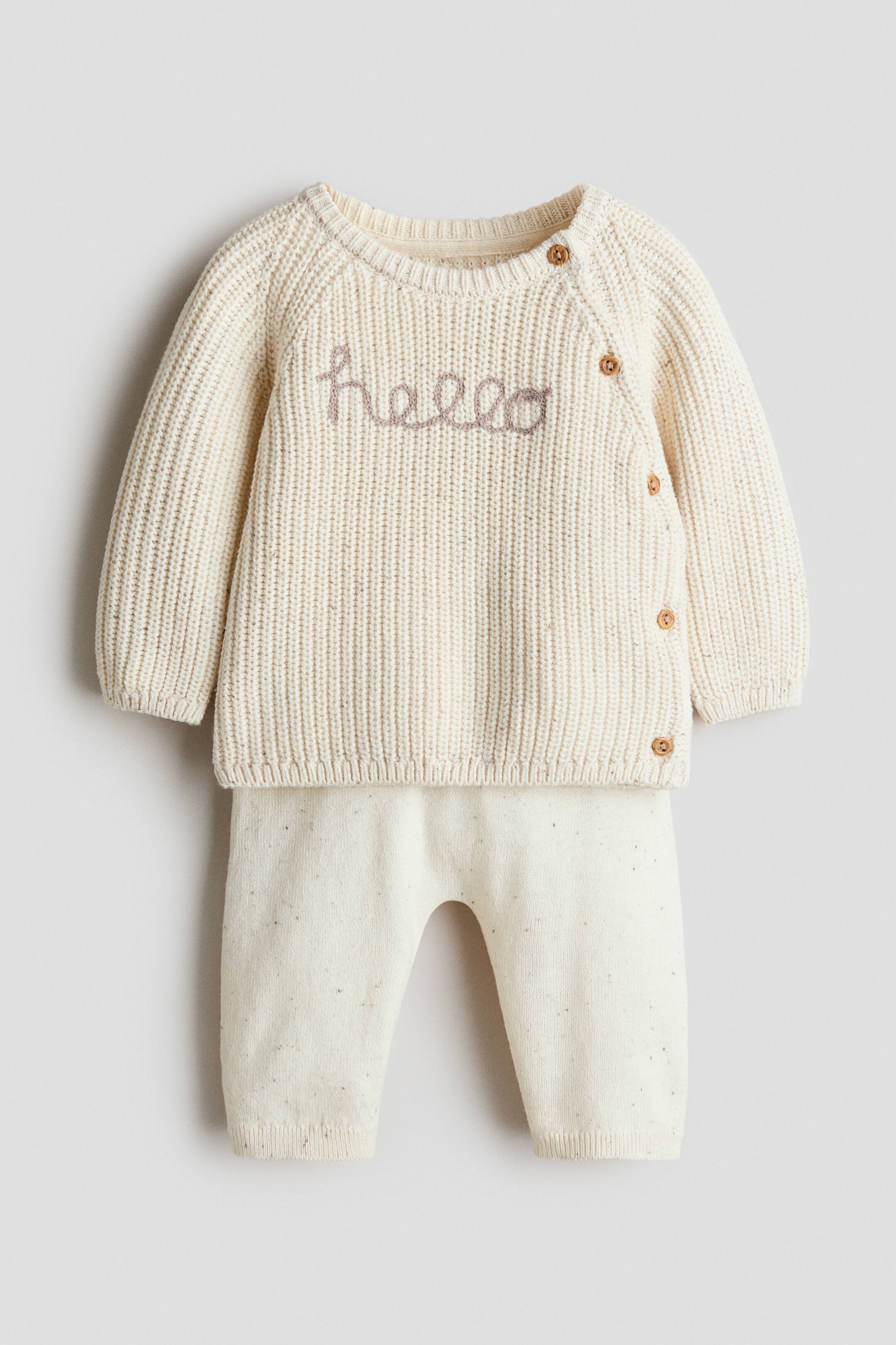 View larger image: The product features a cream fine-knit cotton set, shown from the front and comprising a jumper and matching trousers. The jumper showcases a ribbed texture with light brown "hello" embroidery on the front, long raglan sleeves, and a side opening with wooden-look buttons, finished with ribbed cuffs and hem. The matching trousers feature a covered elasticated waist, ribbed hems, and a subtle speckled texture.