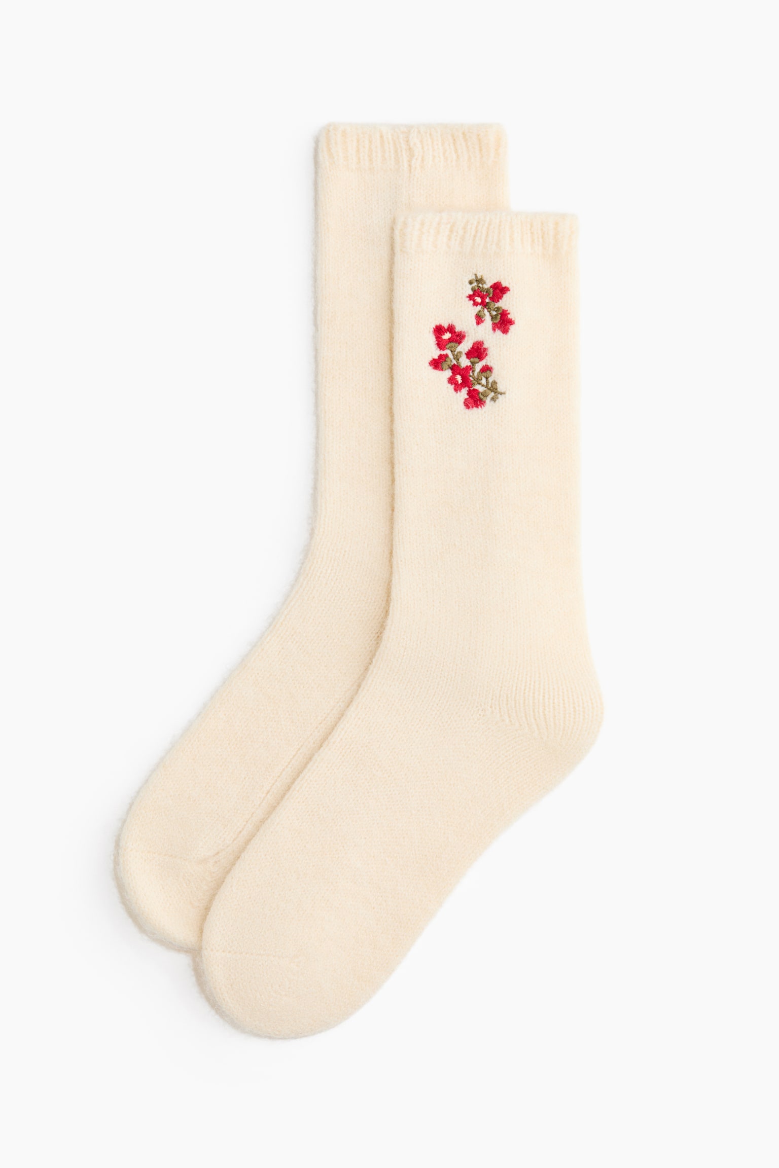 Ein Paar cremefarbene Stricksocken liegt flach und zeichnet sich durch eine weiche Struktur sowie gerippte BÃ¼ndchen aus. Die vordere Socke zeigt oberhalb des KnÃ¶chels ein kleines, gesticktes Blumendesign mit roten BlÃ¼ten und grÃ¼nen Stielen.
