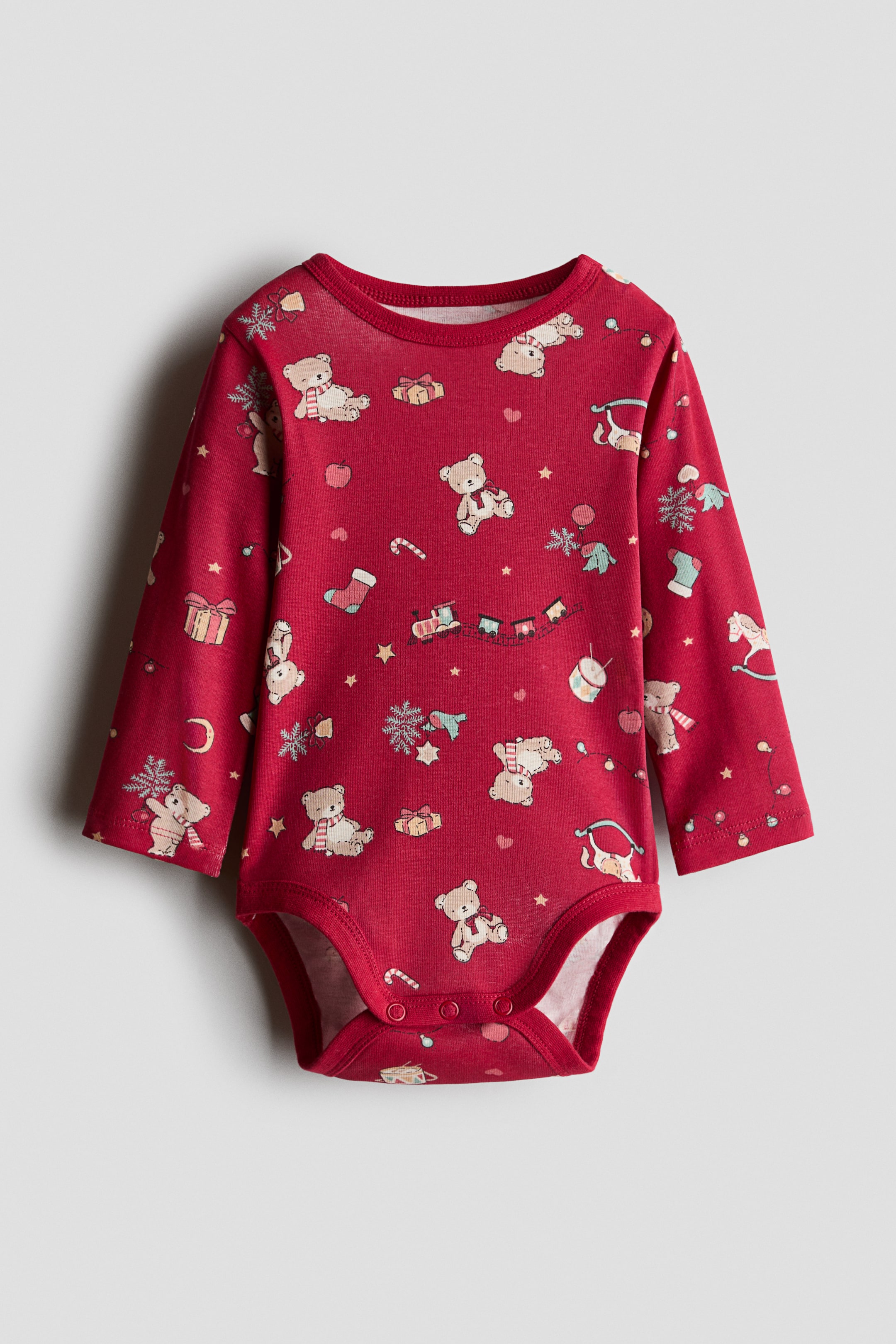 View larger image: A red, long-sleeved baby bodysuit features a festive print of teddy bears, toy trains and stockings. The front view highlights a round neckline with ribbed trim and press-stud fastenings at the bottom.