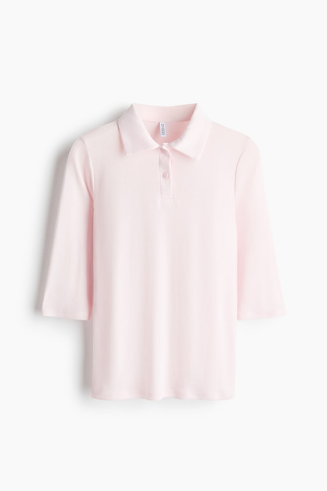 A light pink polo shirt in fine-ribbed jersey is shown front-facing, featuring a classic collar, a two-button placket, and three-quarter-length sleeves. The shirt is designed with a straight hem, presenting a clean and simple silhouette. - 1
