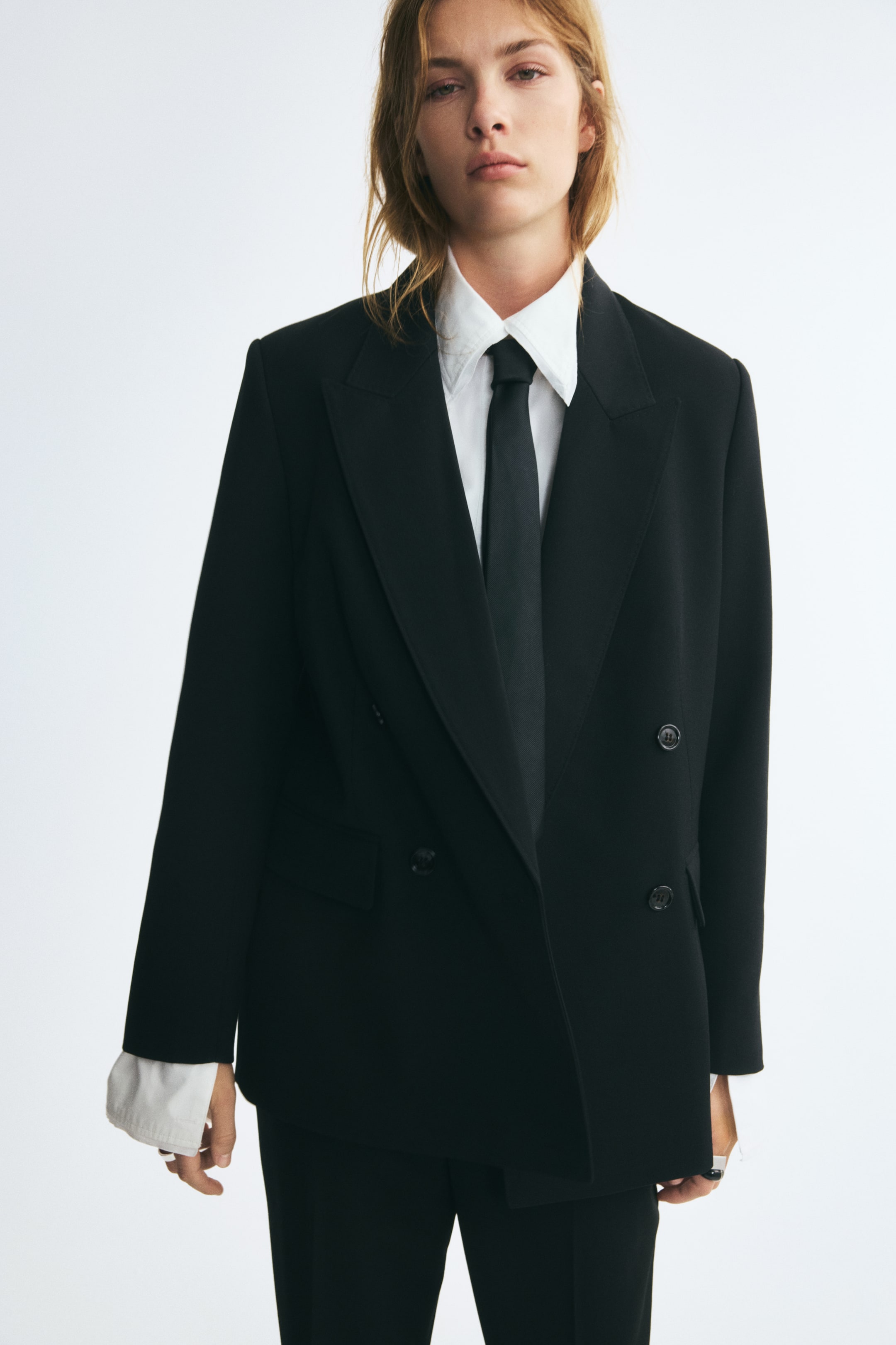 Vis større bilde: En lang, svart dobbeltspent blazer med brede spisse jakkeslag og markerte skulderputer er båret av modellen. Blazeren er stylet over en hvit skjorte med krage, med svart slips, og kombinert med matchende svarte bukser.