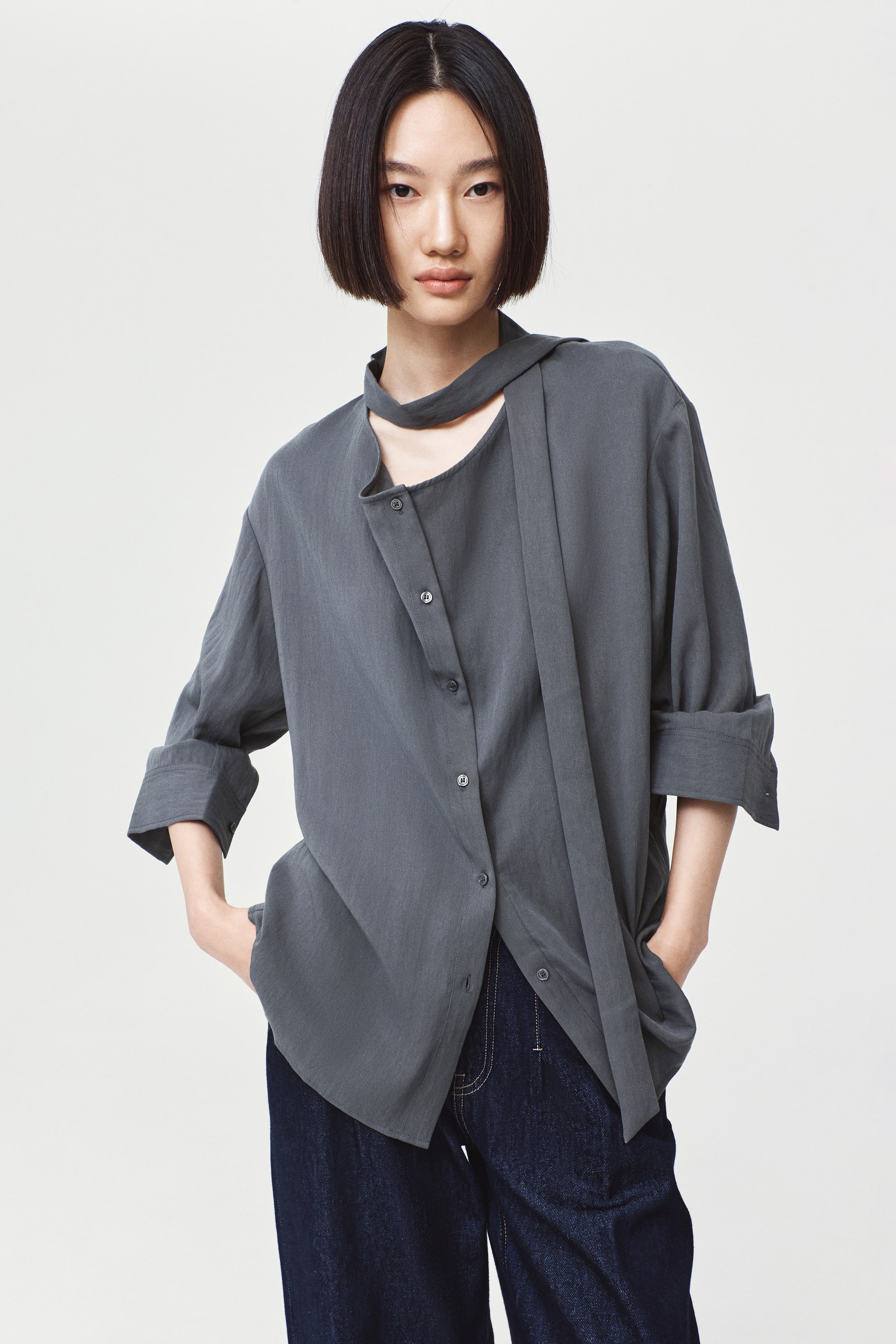 Xem ảnh lớn hơn: Người mẫu đang mặc một chiếc áo blouse dáng rộng, màu xám than, có phần cài nút phía trước, vai trễ và cổ drape bất đối xứng. Chiếc áo được phối cùng quần jeans denim tối màu, với người mẫu đặt tay trong túi quần.