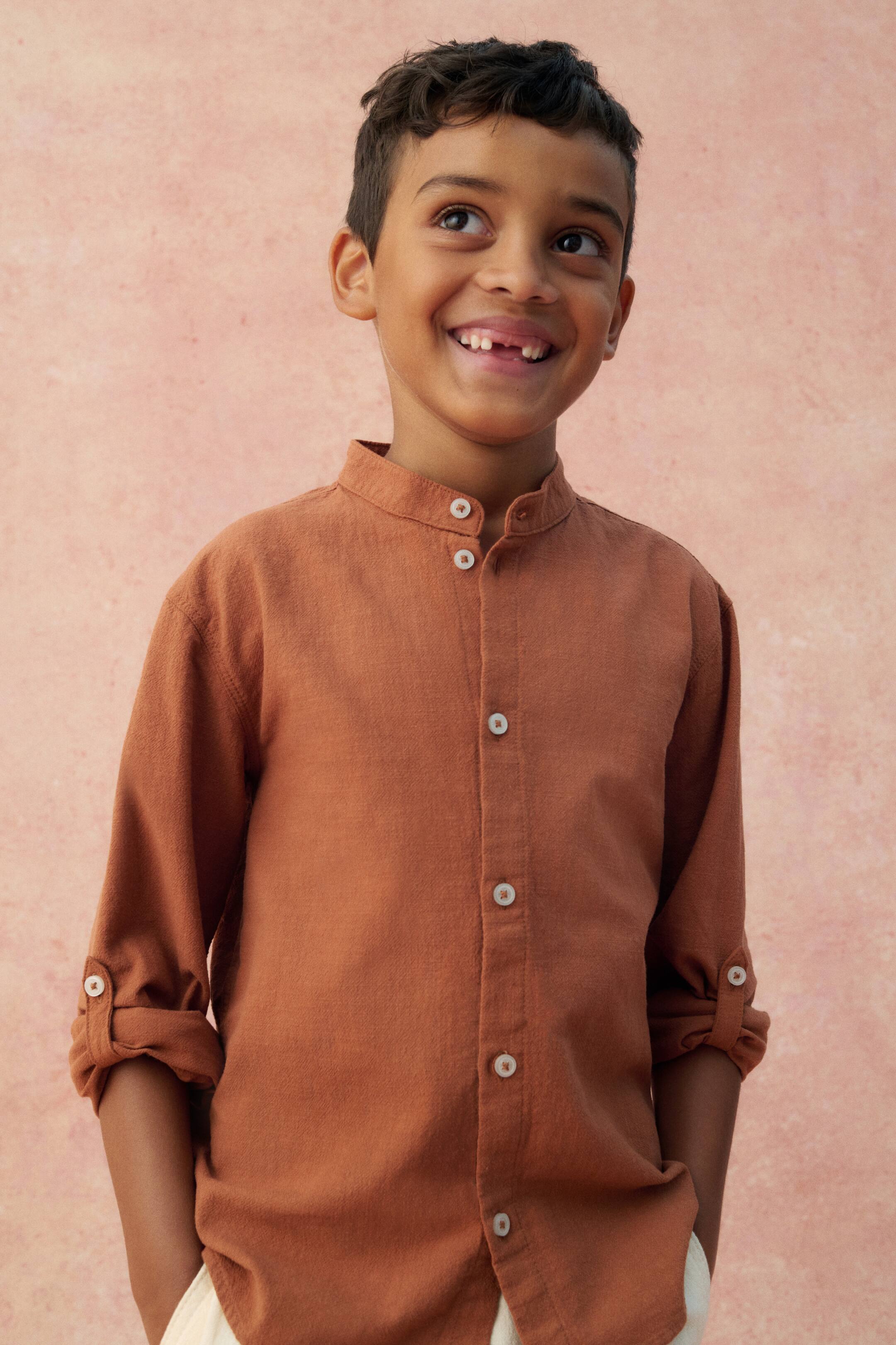 View larger image: A rust-colored button-down shirt, made from a subtly textured fabric, is being presented from a slightly angled front view. It features a band collar and light-colored buttons running down its front placket, with its long sleeves rolled up and secured by buttoned tabs.